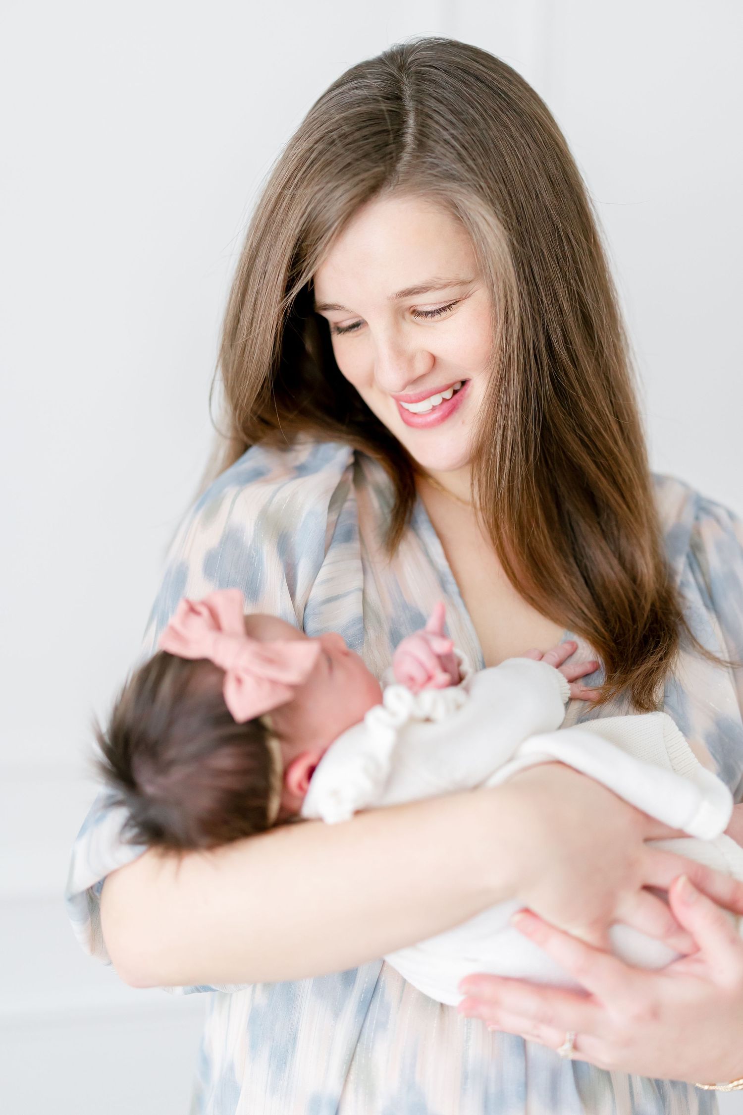 A mother in a blue and white dress smiles lovingly while cradling her newborn baby against a light background.