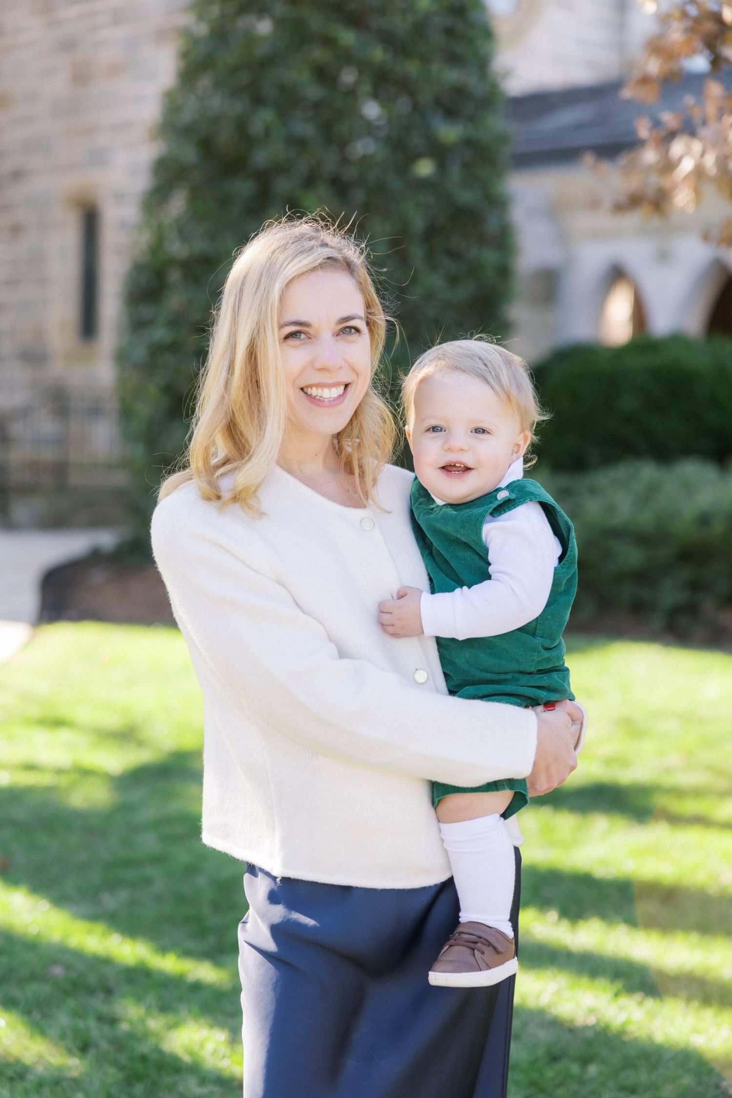 A person in a white sweater holds a smiling toddler wearing green and white on a sunny day outside a stone building.