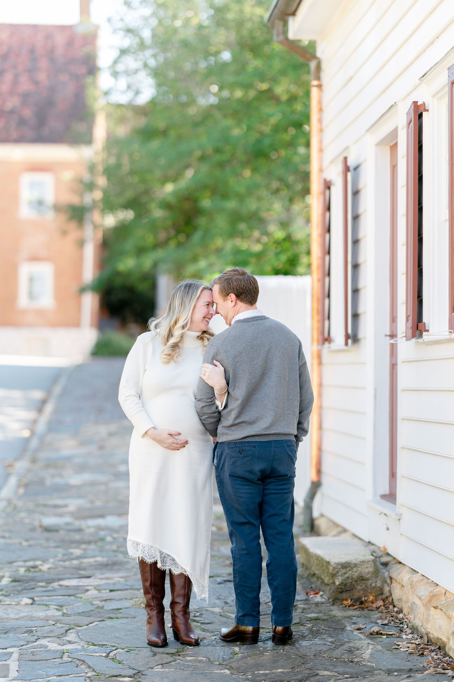 Expecting couple standing forehead to forehead on a cobblestone street beside a white historic building during a maternity photo session.