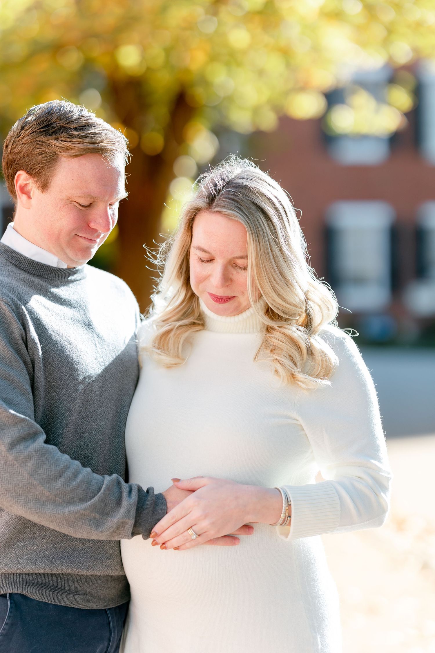 Smiling expectant couple in cozy sweaters gently cradling the woman's baby bump outdoors beneath golden autumn trees.