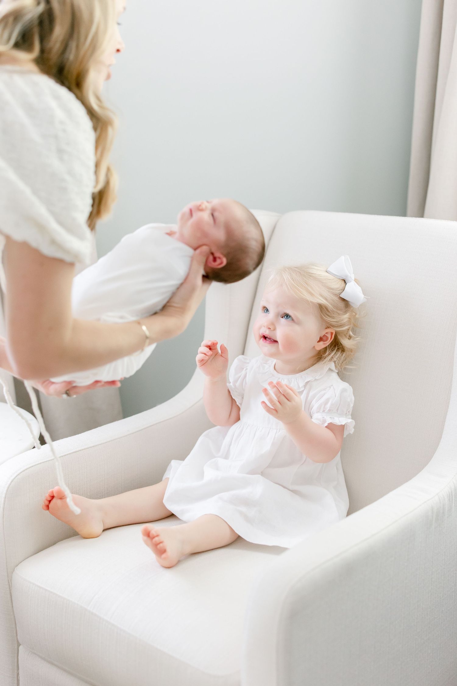 A toddler in white dress sits in chair clapping while looking at baby.