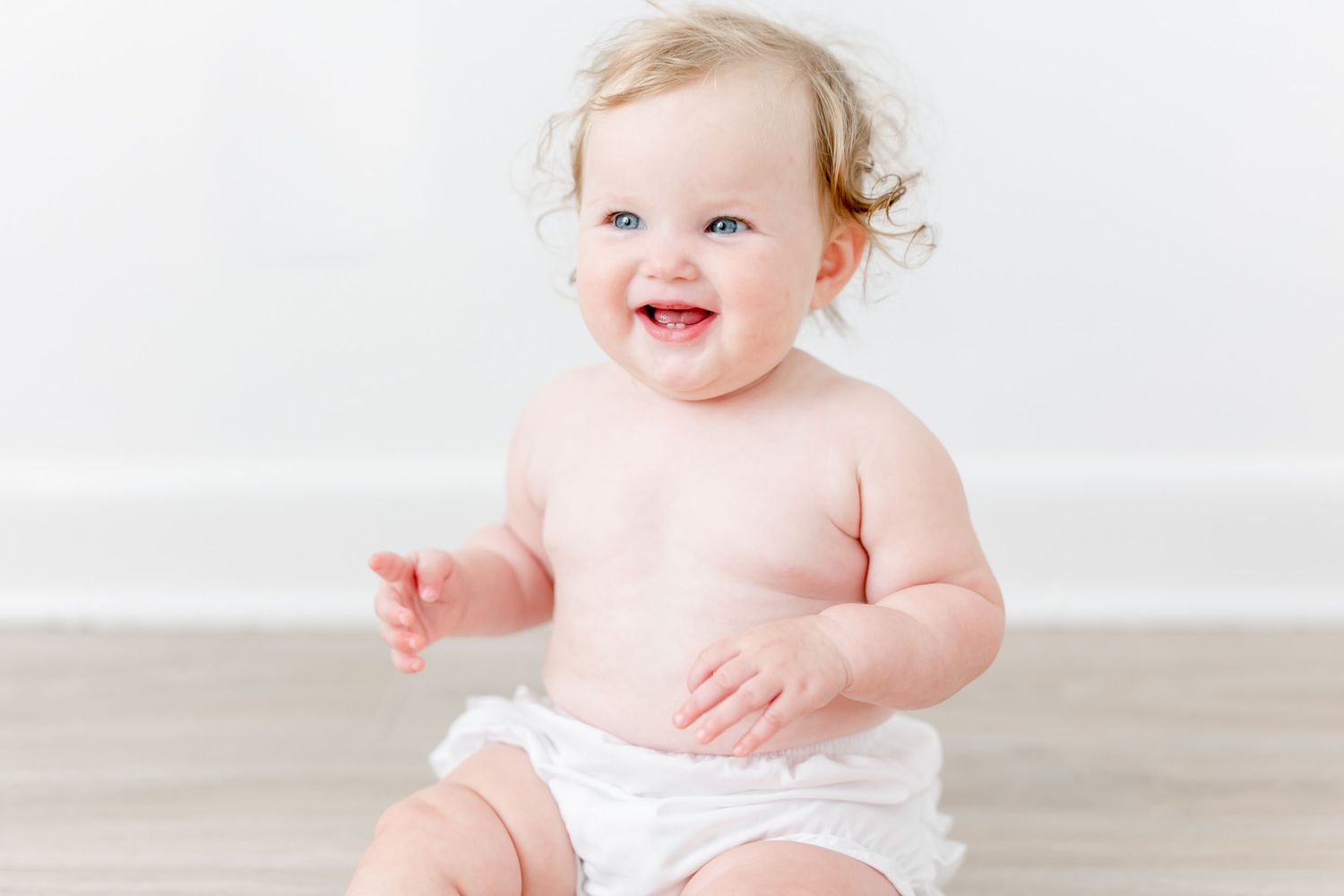 Happy baby wearing white diaper sits and smiles in front of white backdrop during photography session.