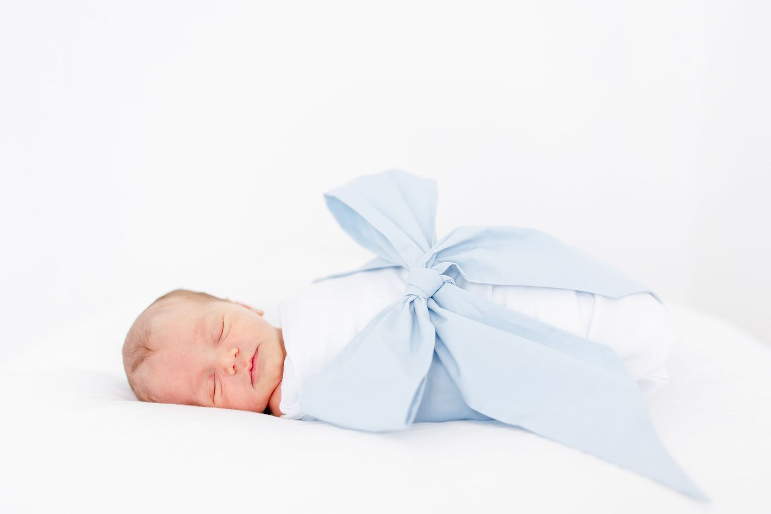 Small baby lies peacefully sleeping in white bedding wearing light blue outfit with matching bow.