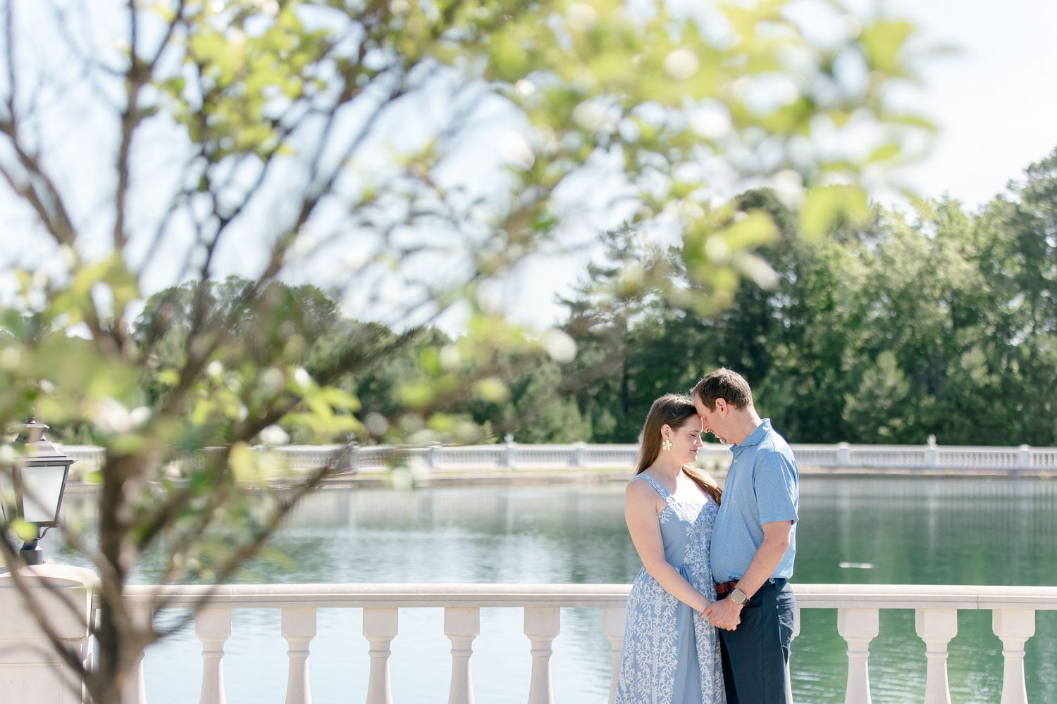 A couple shares a tender moment along a white balustrade overlooking a peaceful pond on a sunny day.