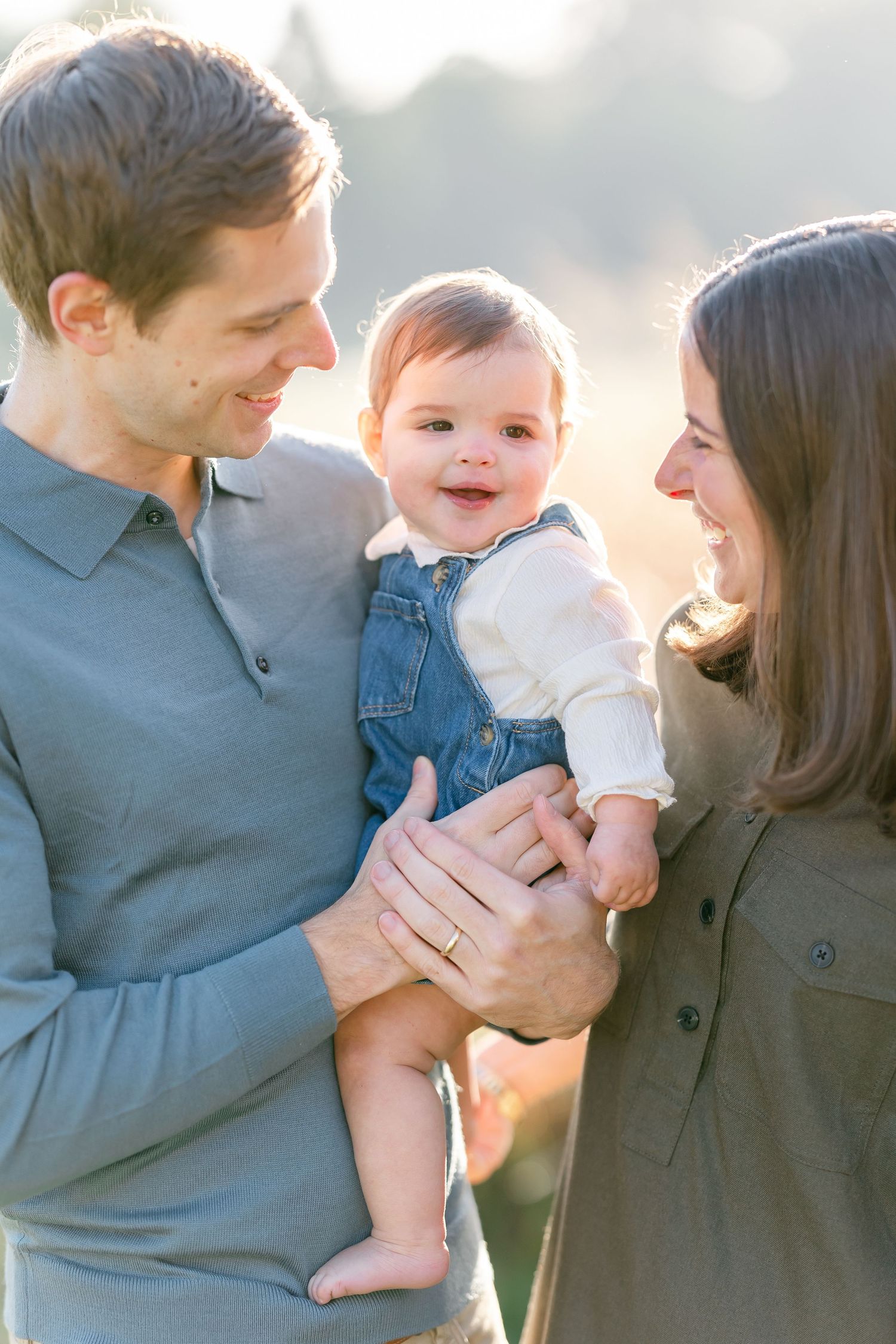 Happy baby in denim overalls smiling at parents outdoors in warm sunlight during a family portrait session.