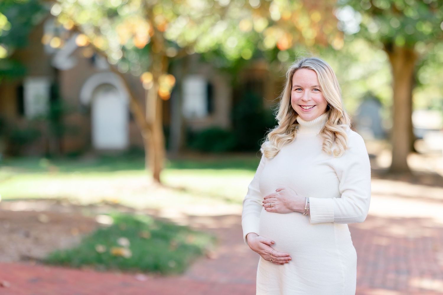 Smiling pregnant woman in a cream knit sweater cradles her baby bump while standing on a brick path near trees and a historic building.