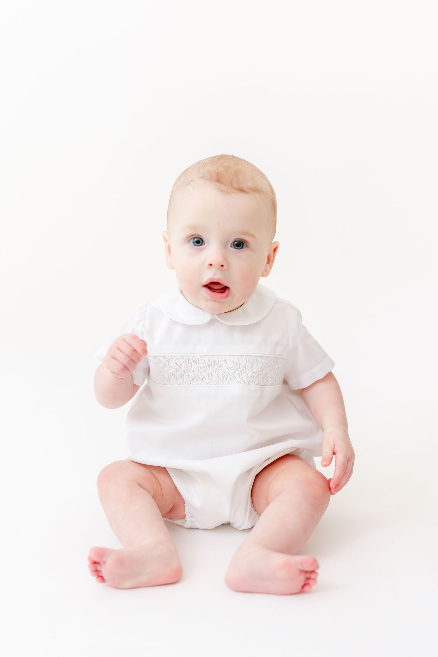 Smiling baby with blue eyes sits in a white smocked romper on a clean white studio background.