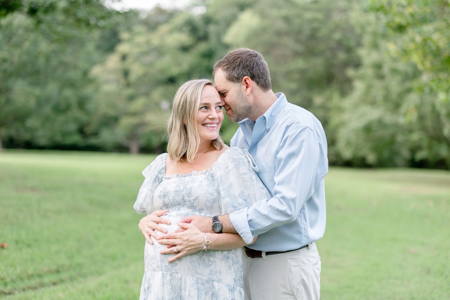 A couple shares a tender moment together on a grassy field with lush green trees in the background.