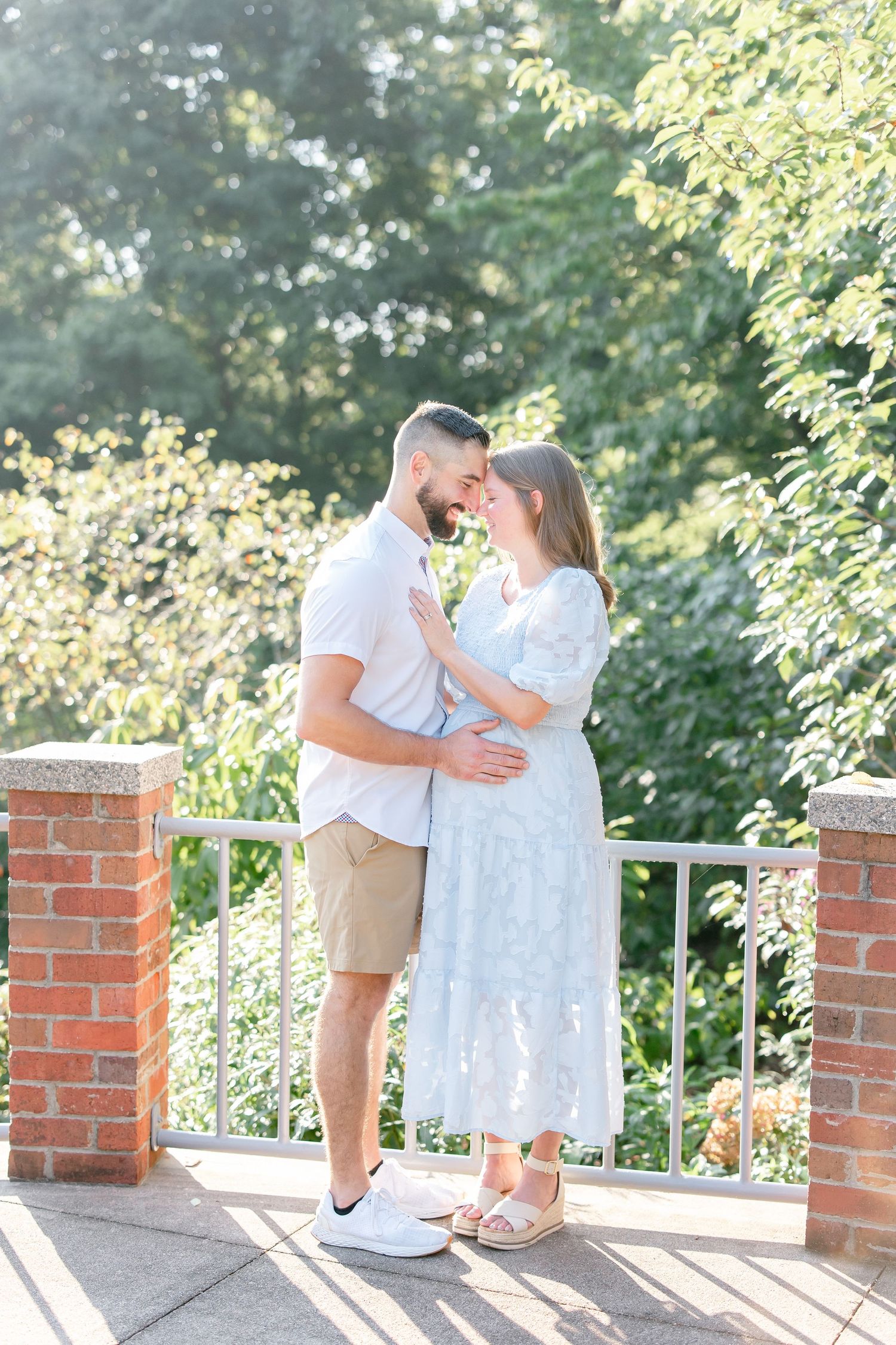 Romantic couple in white and beige outfits share tender moments on a brick-lined porch surrounded by greenery.