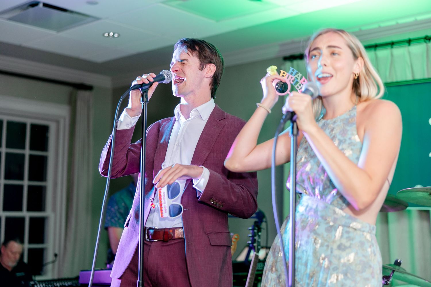 Two performers sing into microphones while performing at an event with green lighting in the background.