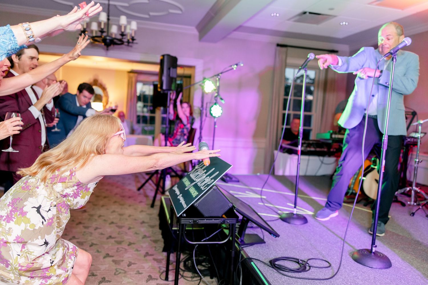 Partygoers perform synchronous dance moves in a venue with purple mood lighting and musical equipment.
