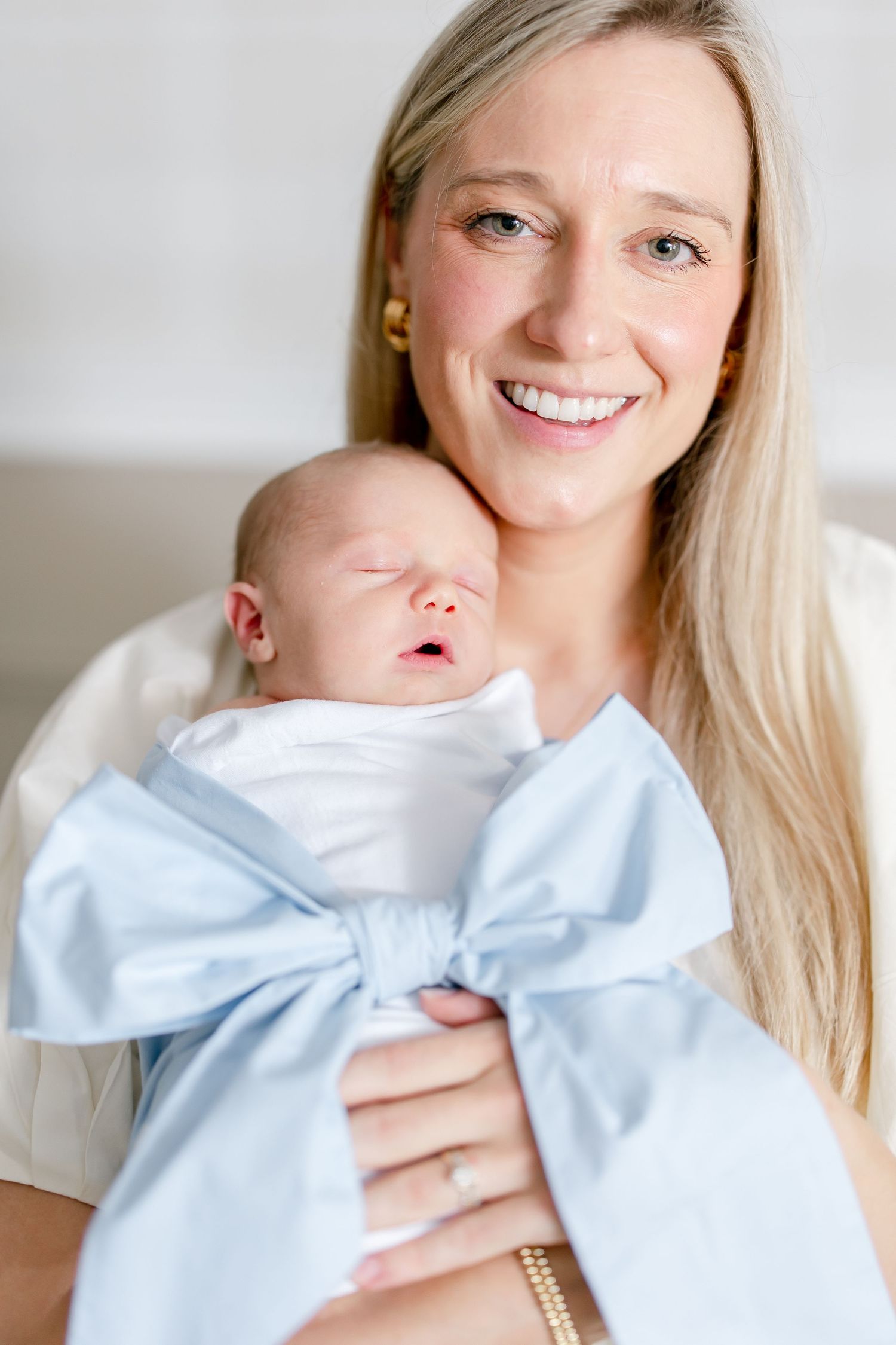 Mother holding newborn baby wrapped in light blue bow while smiling warmly in casual white outfit.