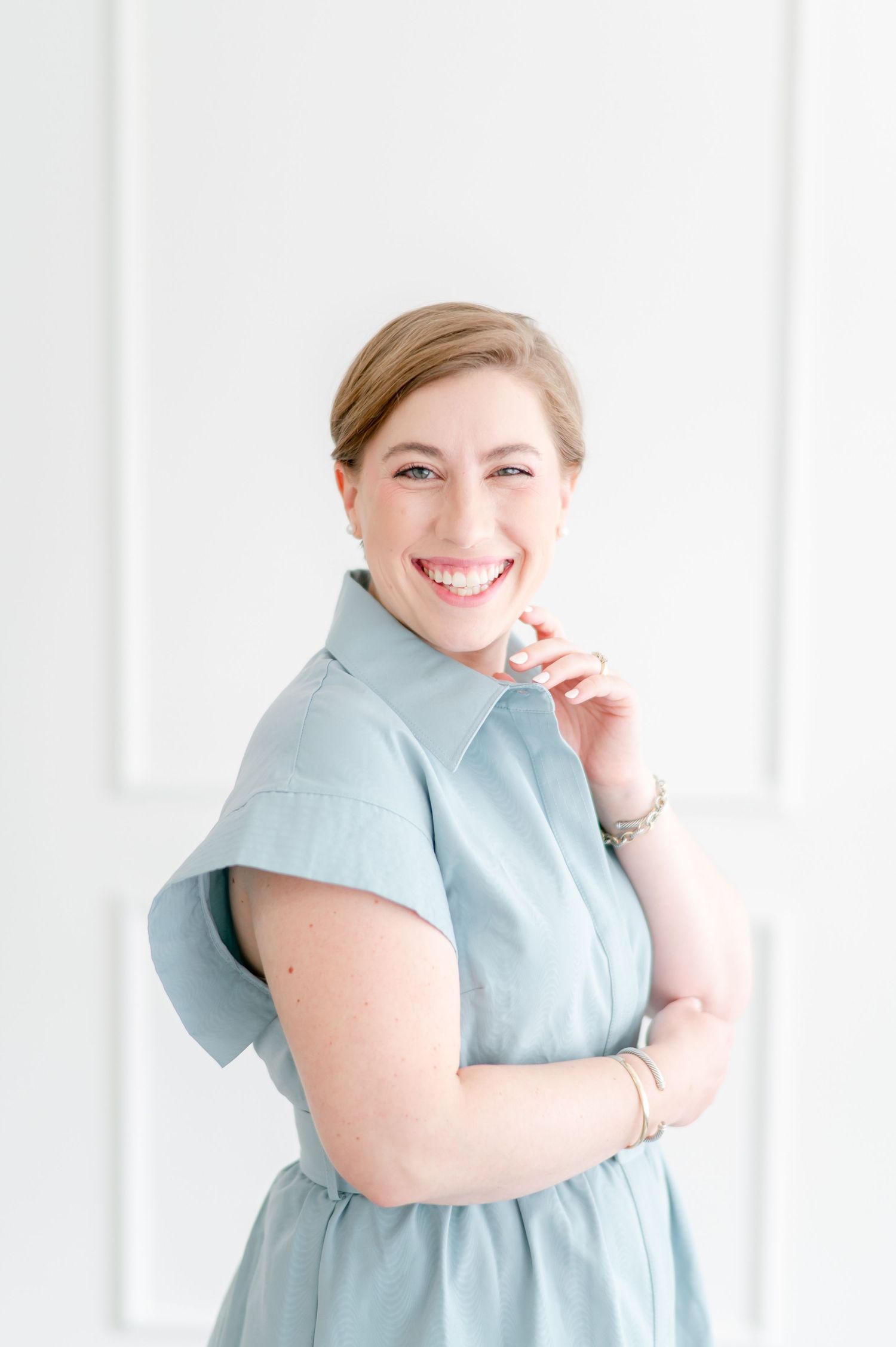 A person in a light blue collared dress smiles warmly while standing against a white wall.