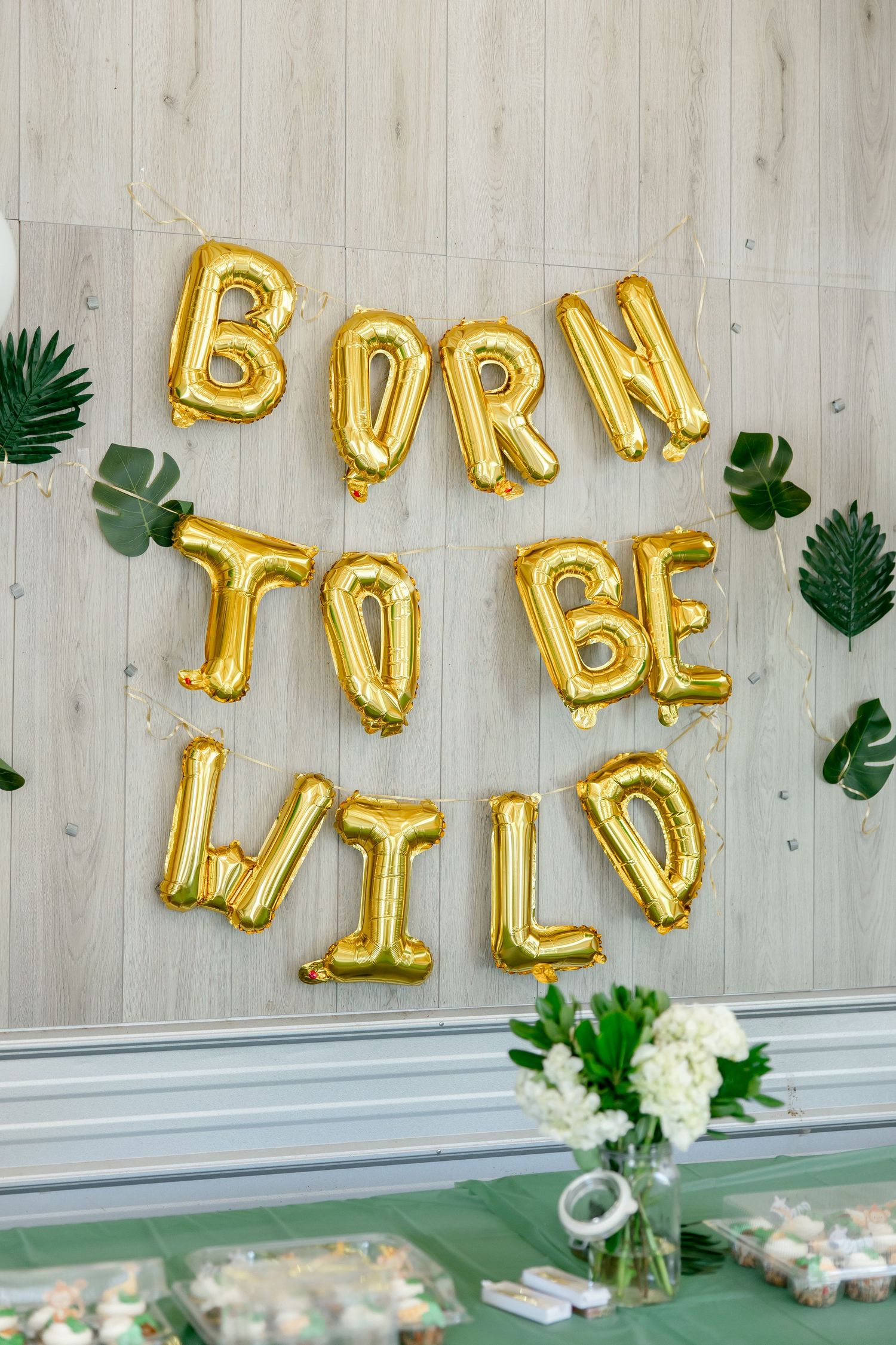 Gold balloon letters spelling 'BORN TO BE WILD' with tropical leaves decoration.