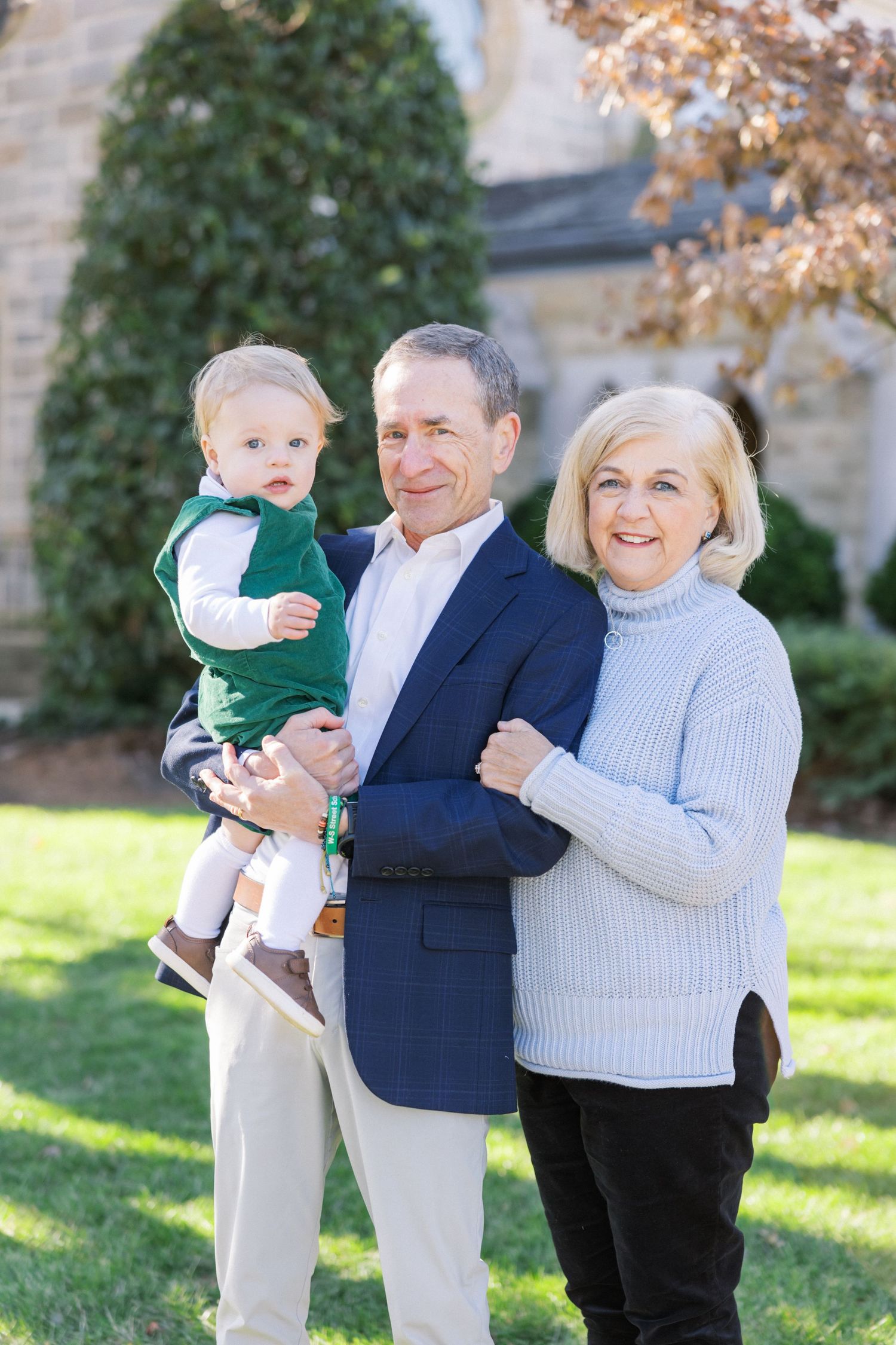 Three people in formal attire pose together outdoors in front of greenery on a sunny autumn day.