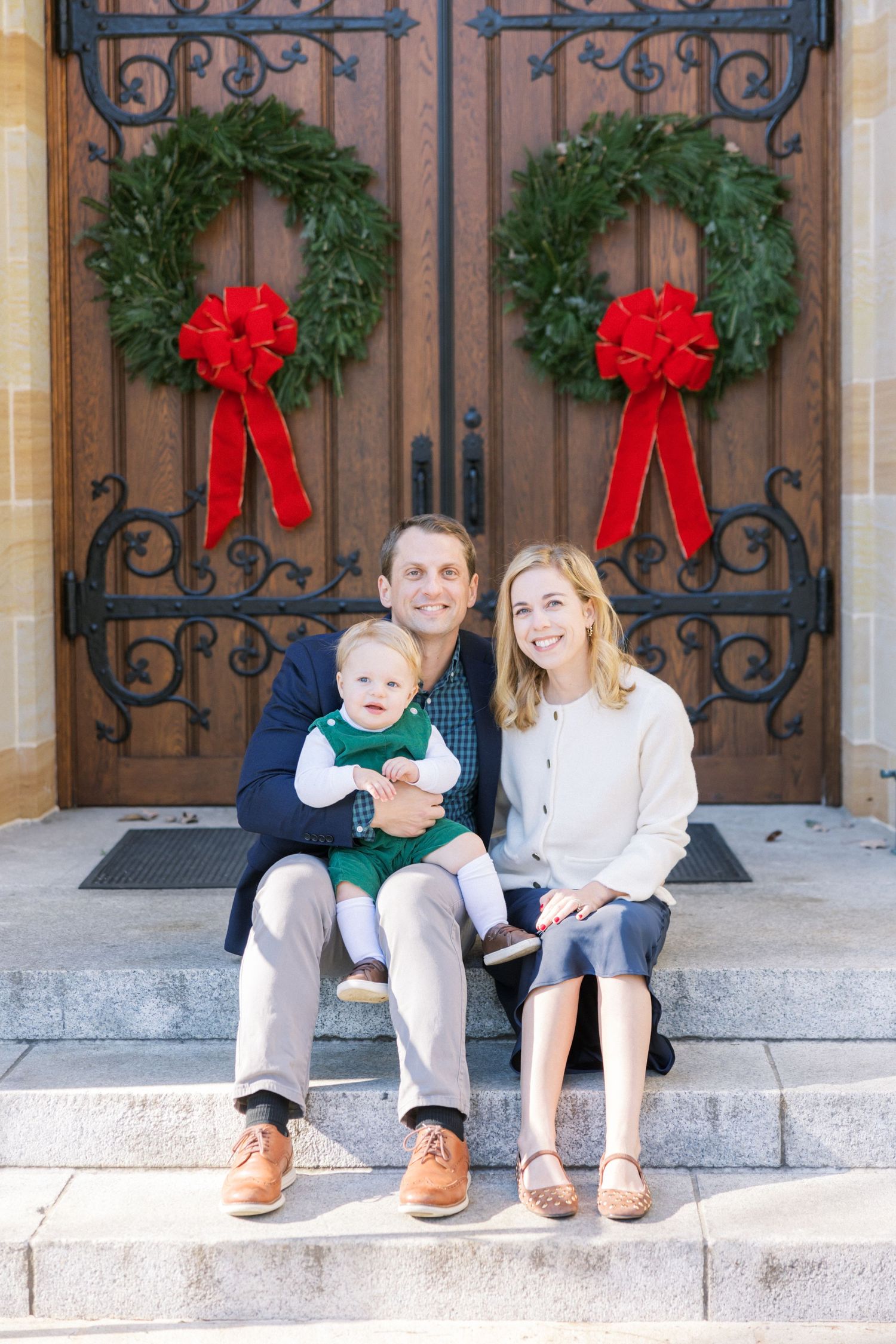 Family sits on church steps with decorative Christmas wreaths and red bows hanging on wooden doors behind them.