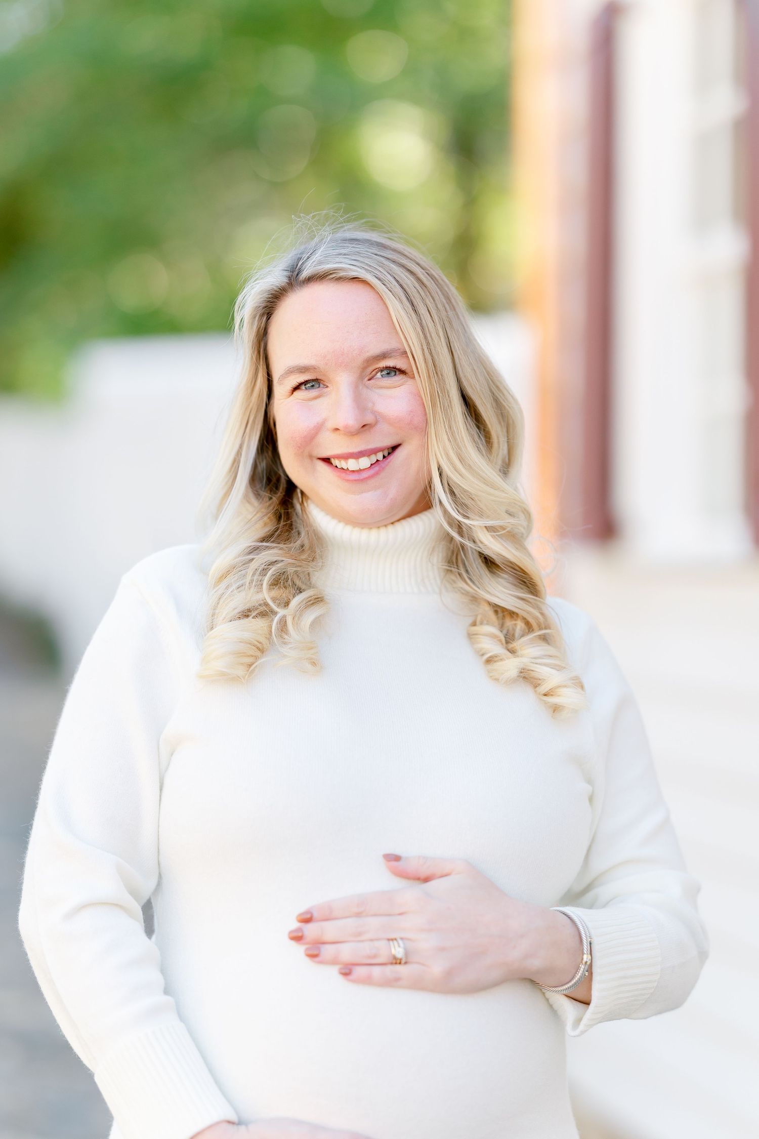 Smiling pregnant woman with long wavy blonde hair in a cream turtleneck sweater gently cradles her baby bump outdoors on a sunny day.