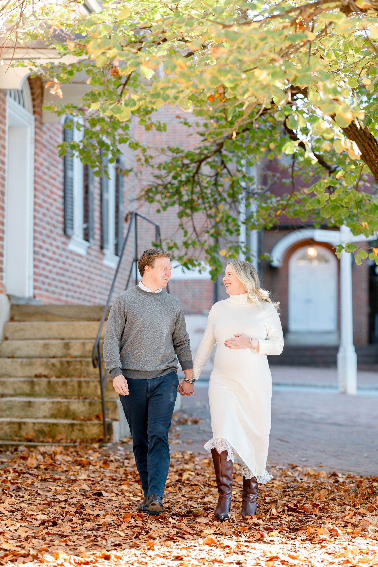 A smiling couple holds hands while walking through fall leaves past a historic brick building, she in a cream dress and boots.
