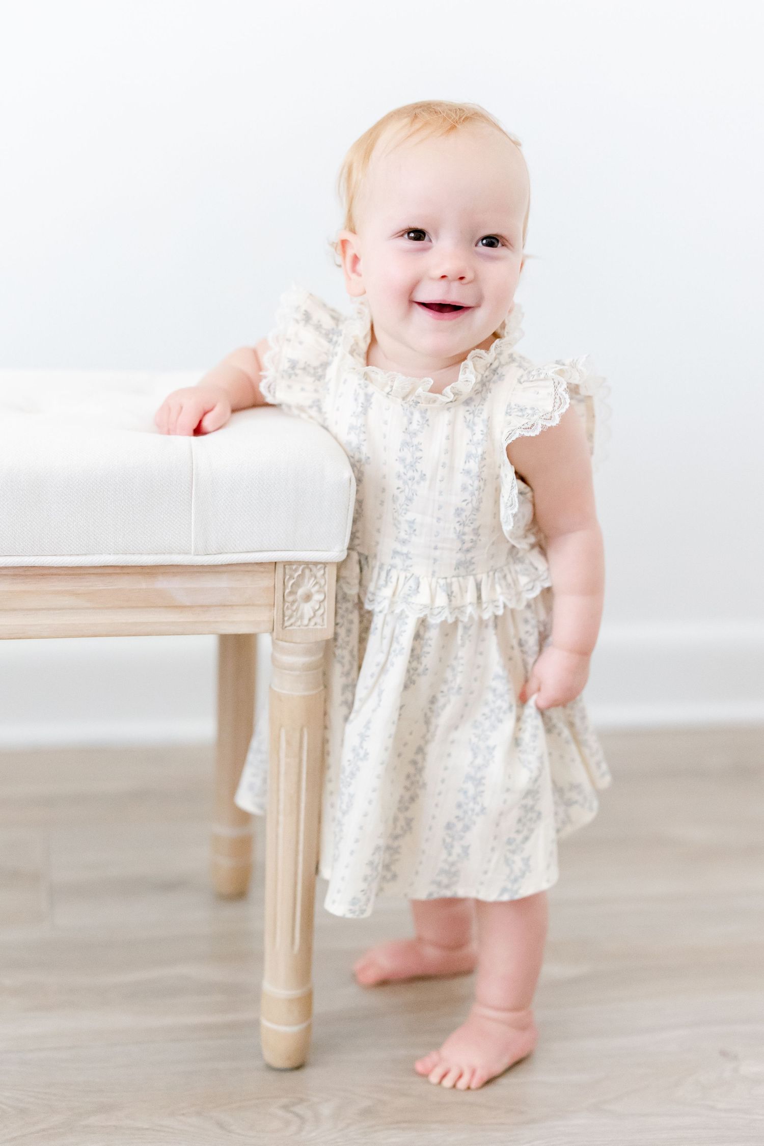 Sweet baby in white lacy dress stands beside wooden table leg on light hardwood floor.