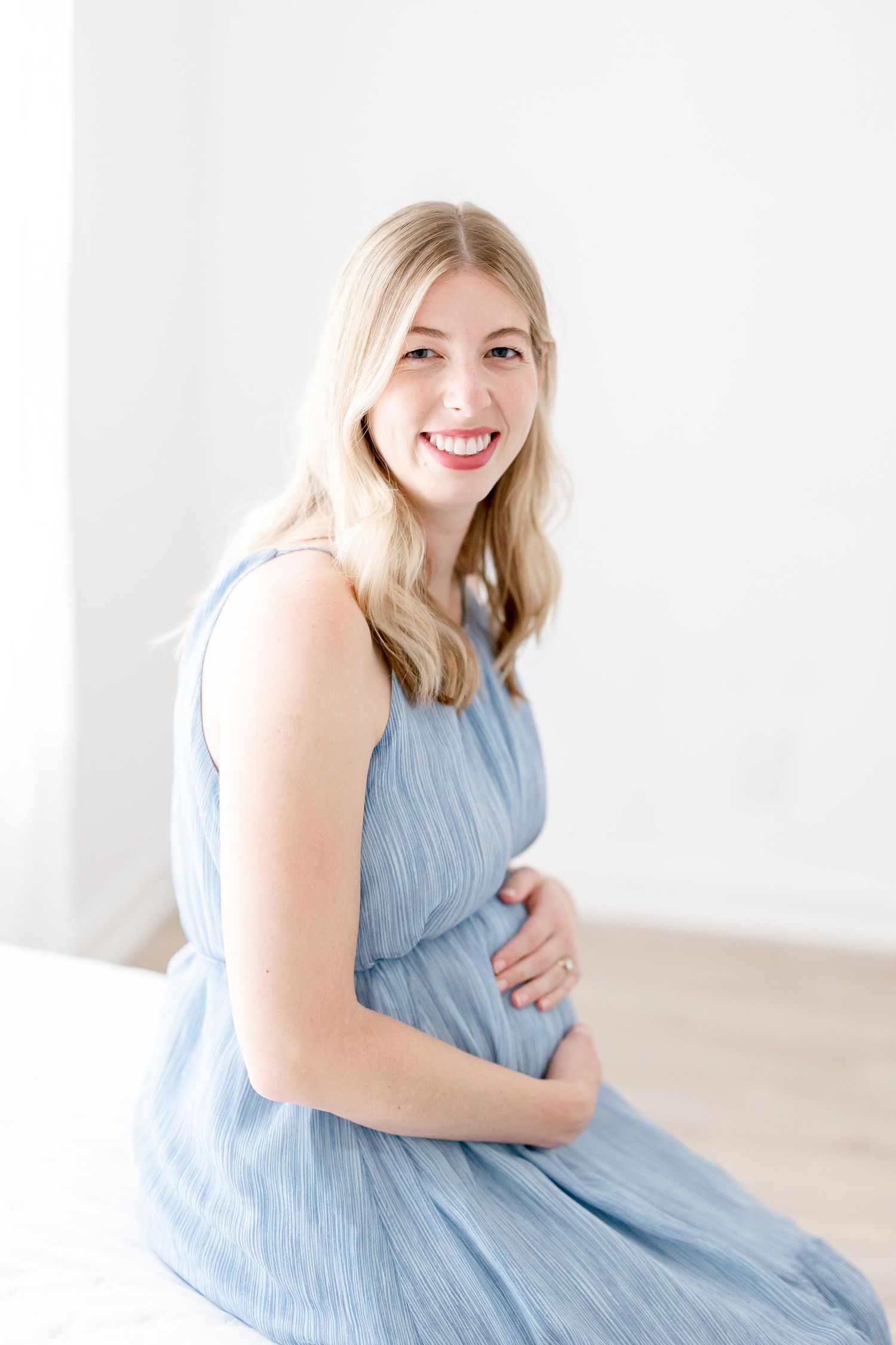 Young woman in a light blue flowy dress poses sweetly against a white backdrop in a minimalist setting.