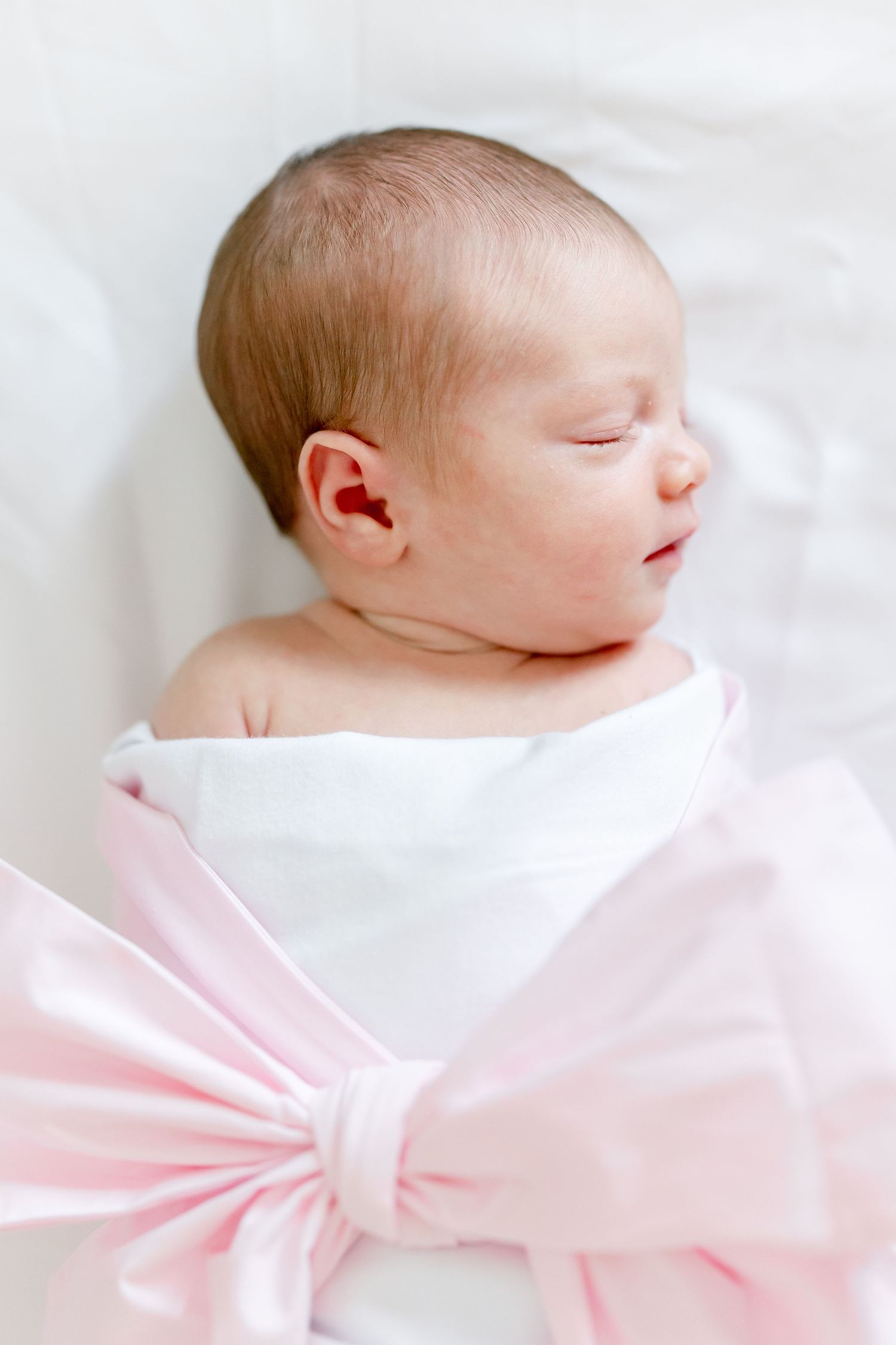 A sleeping newborn swaddled in white and pink fabric against a light background.