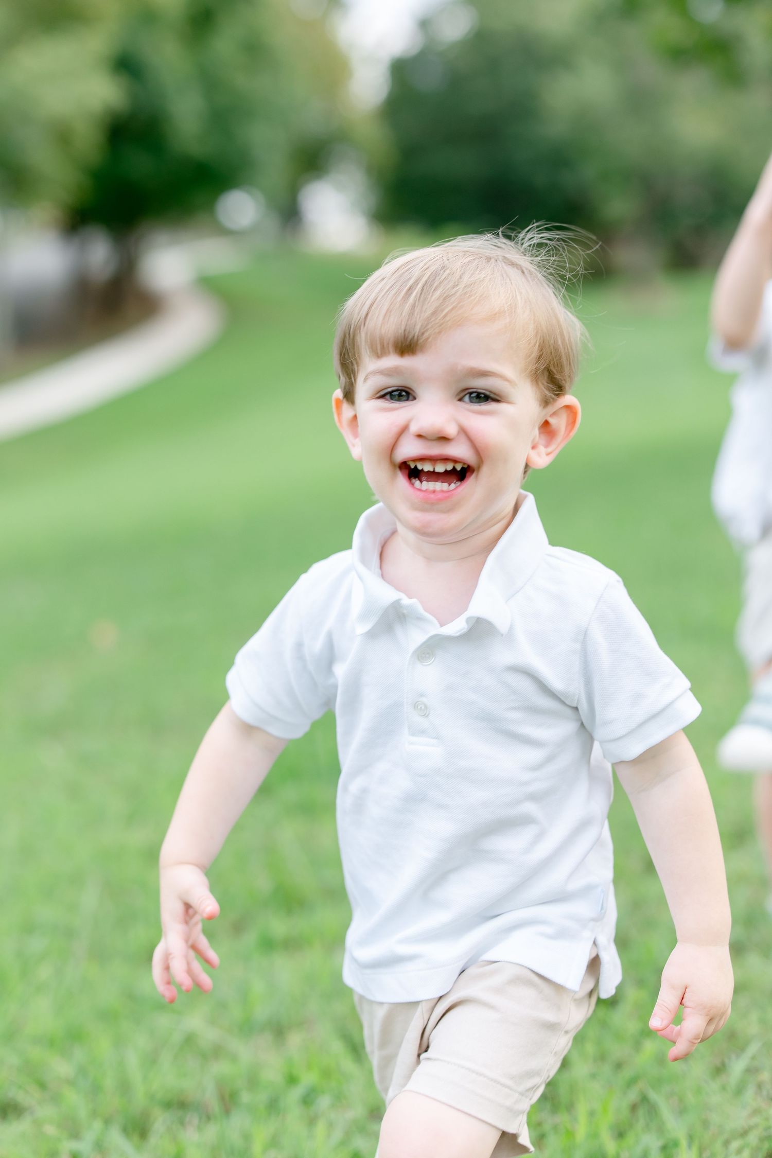 Young toddler in white polo shirt running and laughing joyfully on green grass in a park setting.