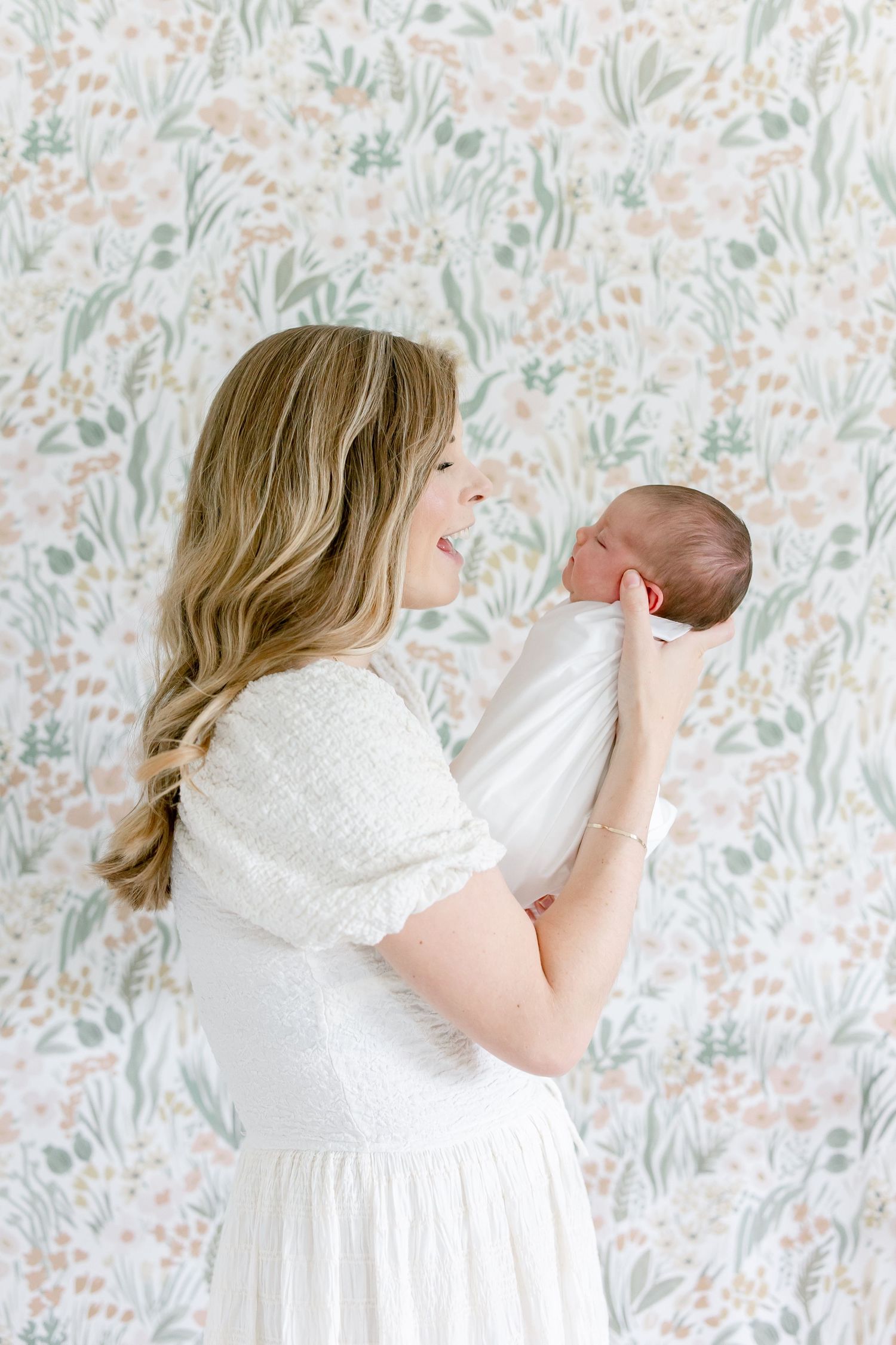 Mother in white dress cuddles newborn against floral wallpaper in light-filled room.