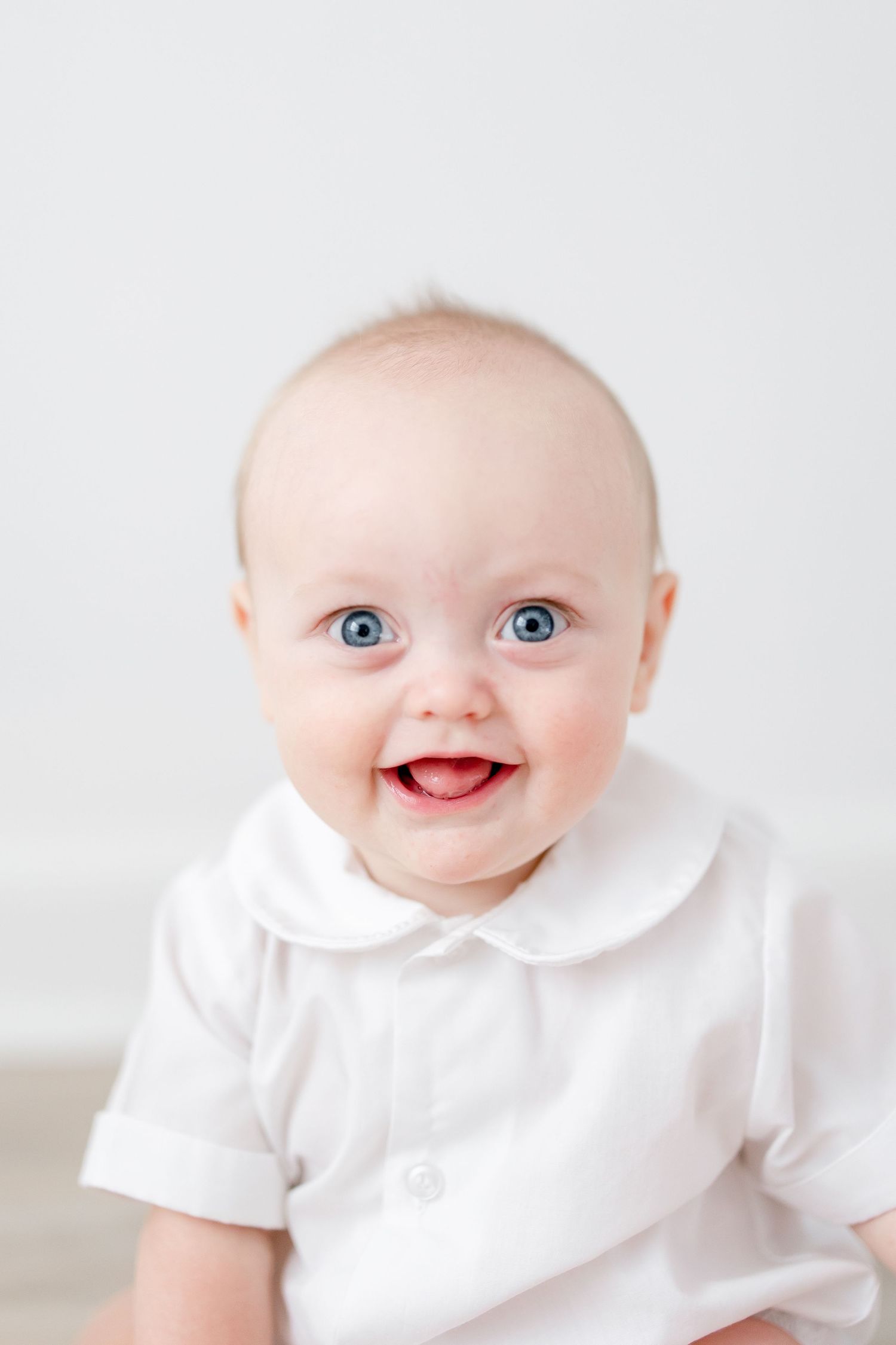 Close-up of smiling baby in white peter pan collar outfit against white background.