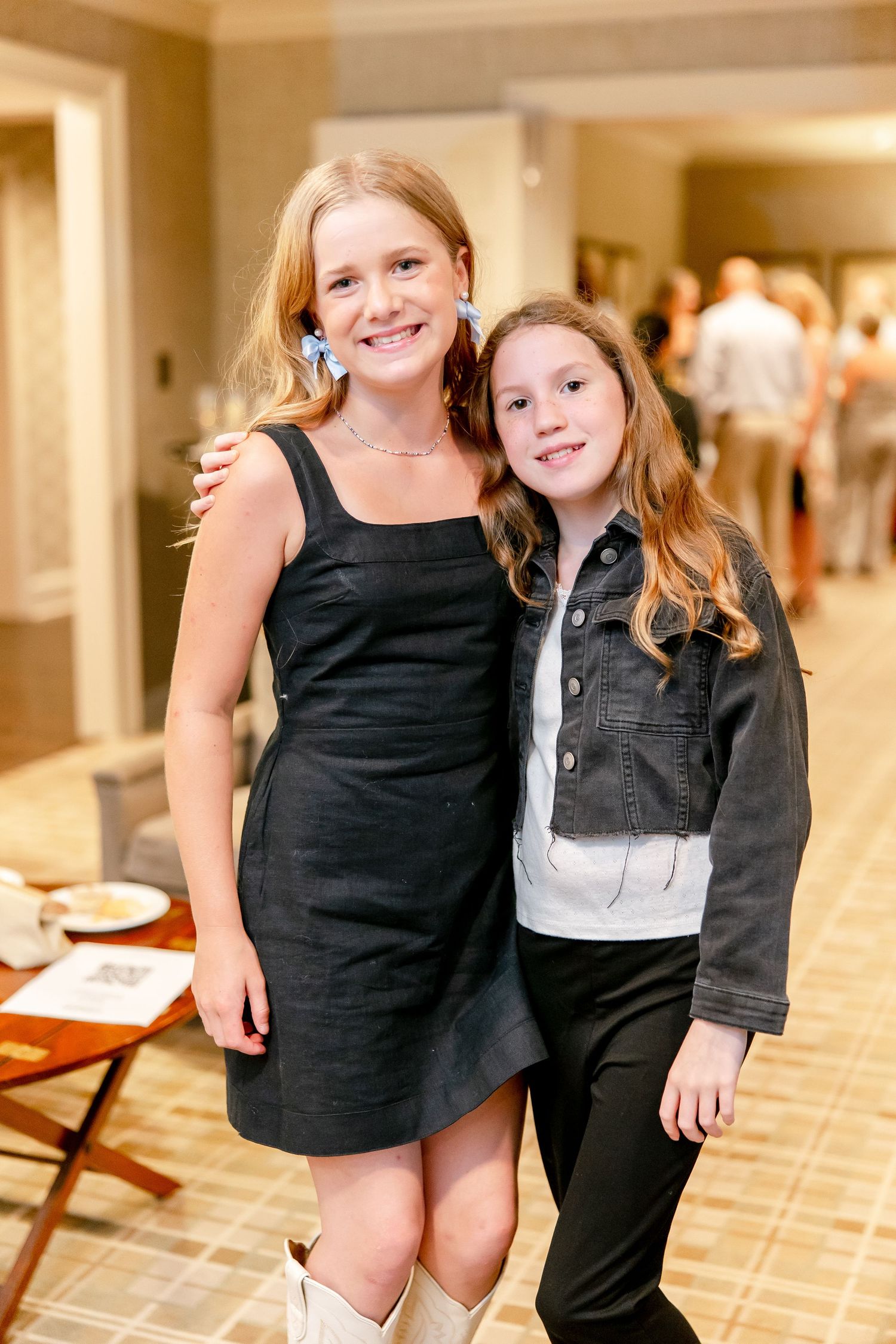 Two young friends in contrasting outfits pose together in a hallway, one wearing a black dress and boots.