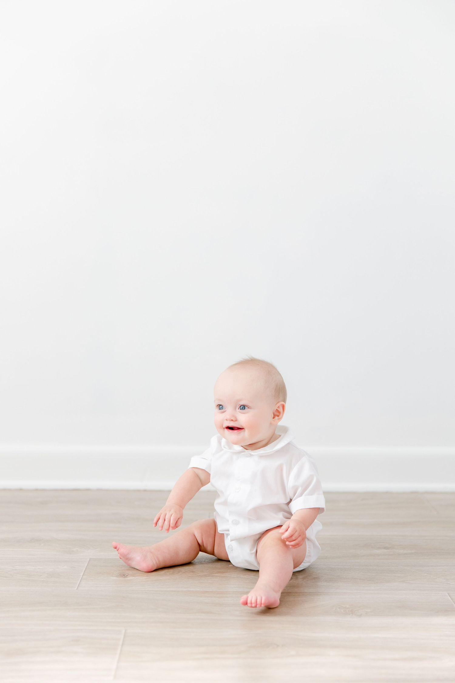 Baby in white onesie sitting on wooden floor during minimalist photo session against white background.