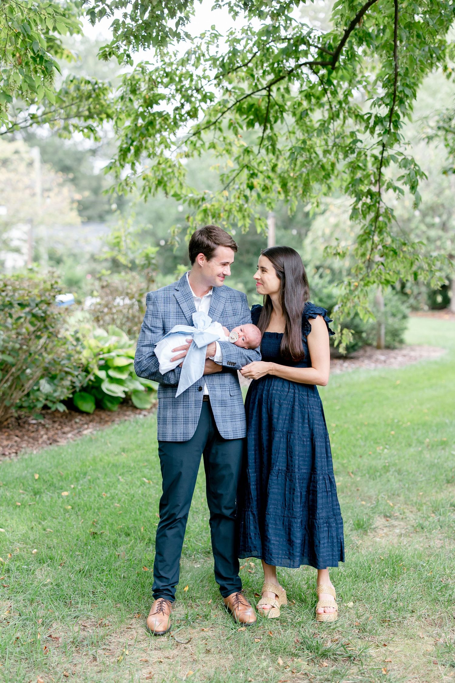 A couple enjoys a sweet outdoor moment together on green grass under leafy trees.