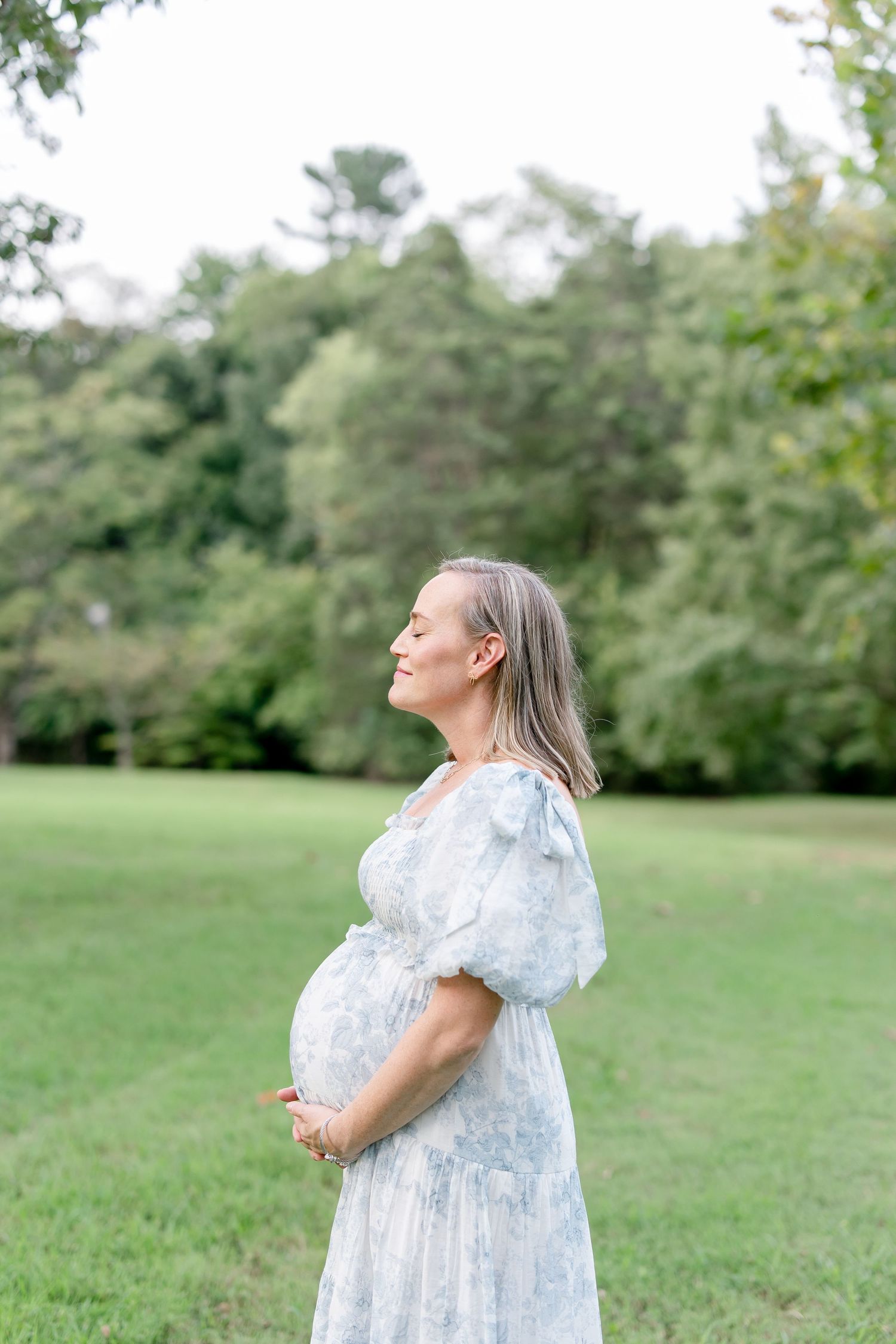 Woman in a flowing light blue dress stands gracefully in a sunlit park.