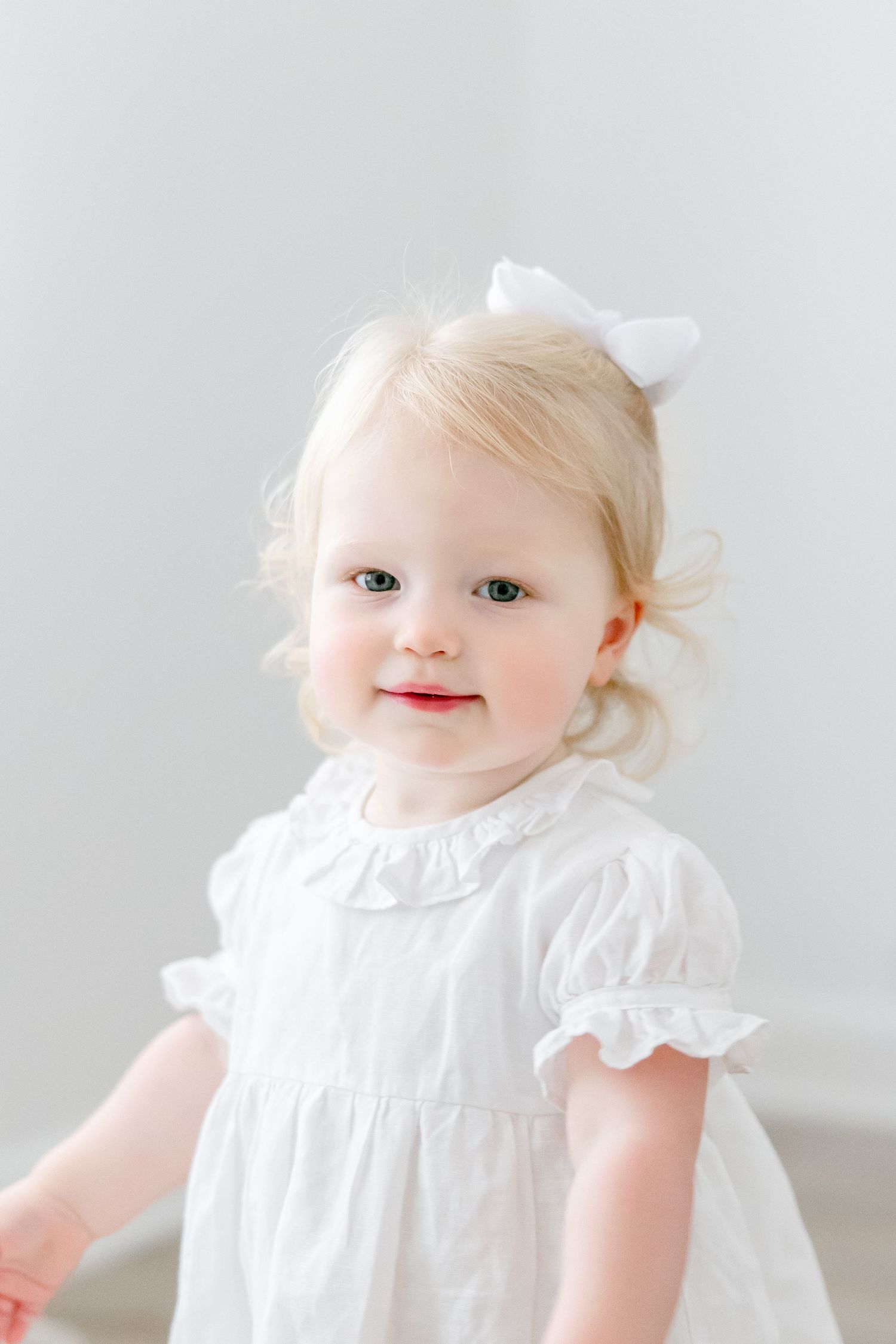 Small child in white dress with bow accessory against minimal white background.