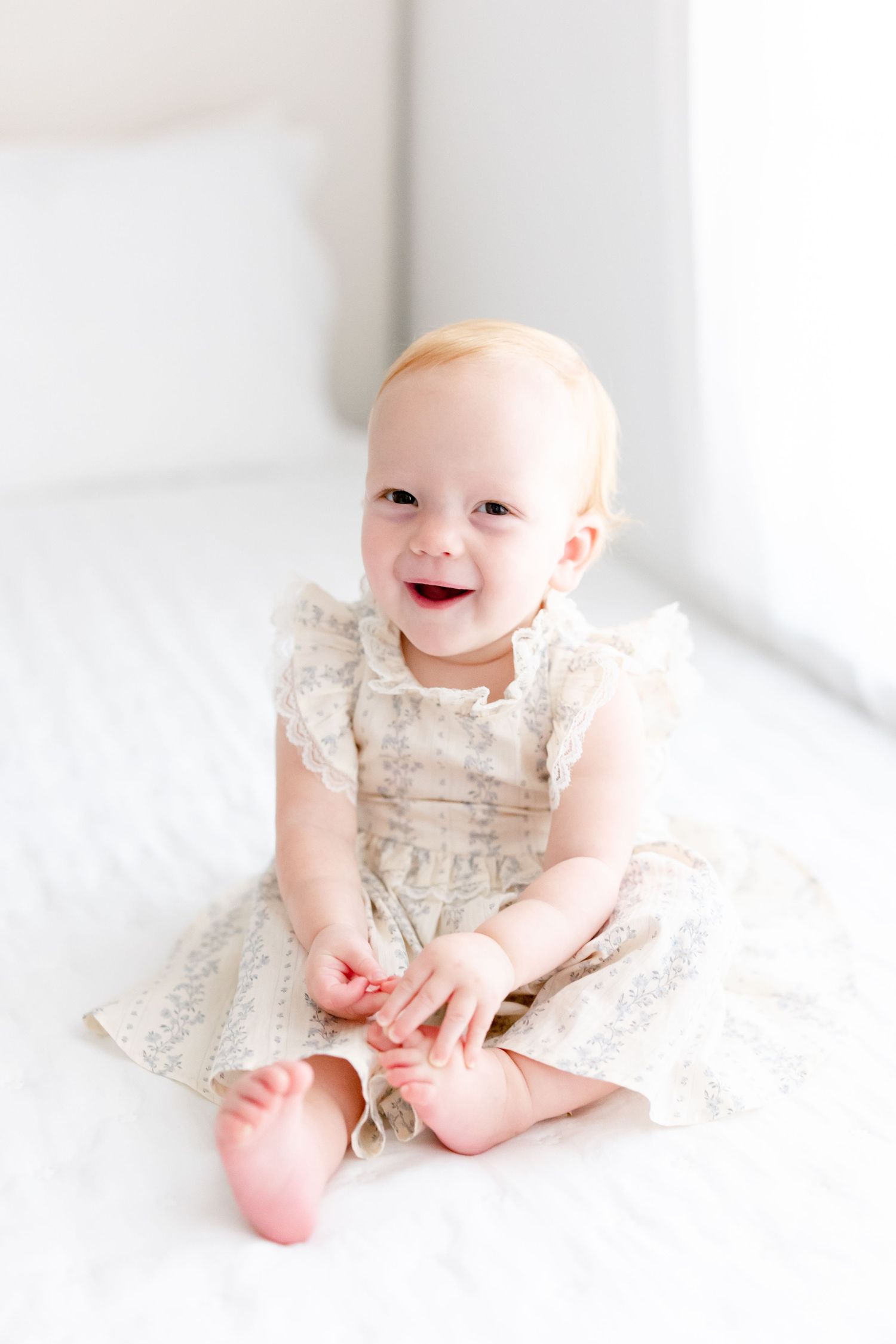 Smiling baby wearing a delicate lace dress sits on white bedding in bright, airy setting.