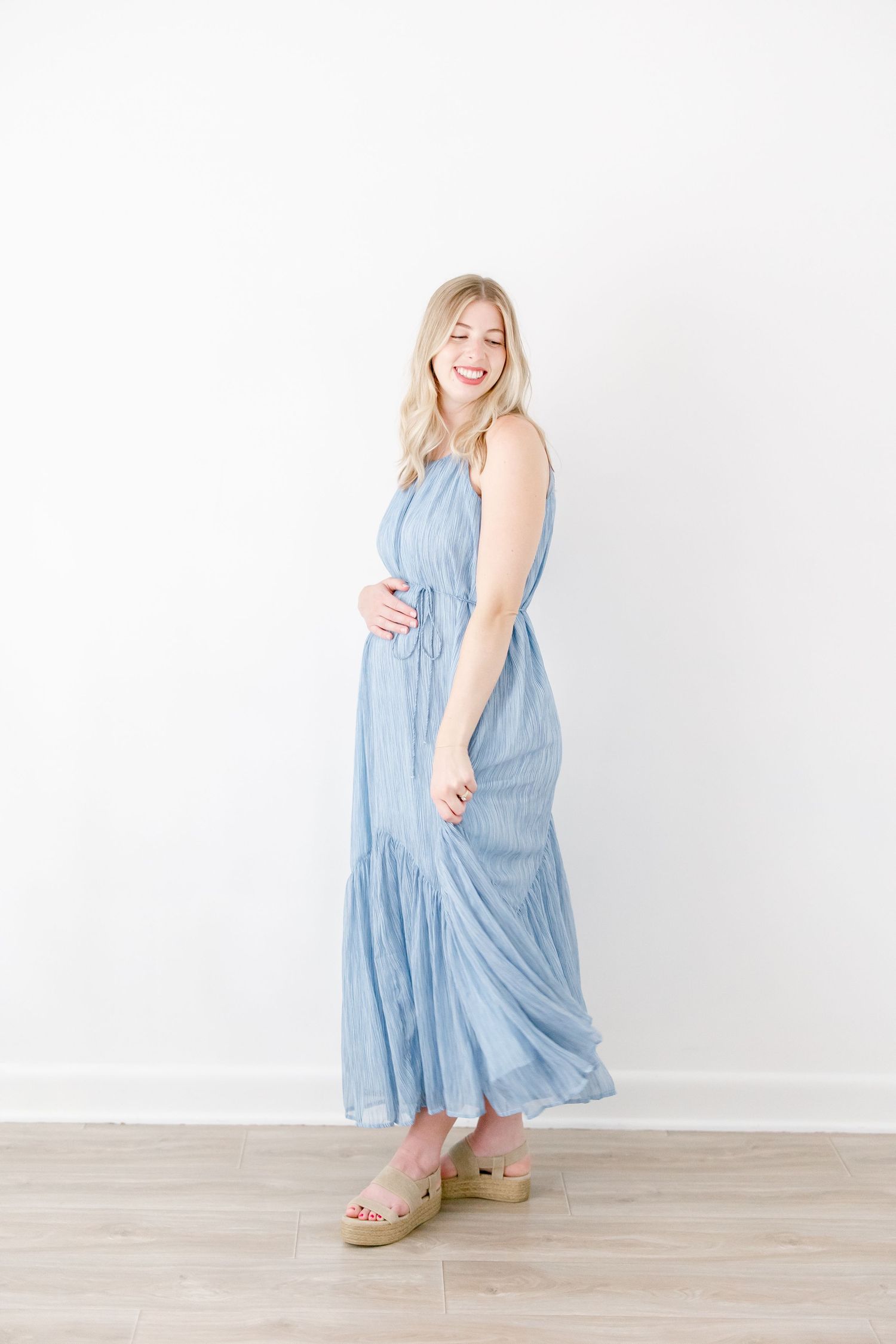 Woman in flowing blue maternity dress twirls and poses playfully against white studio background.