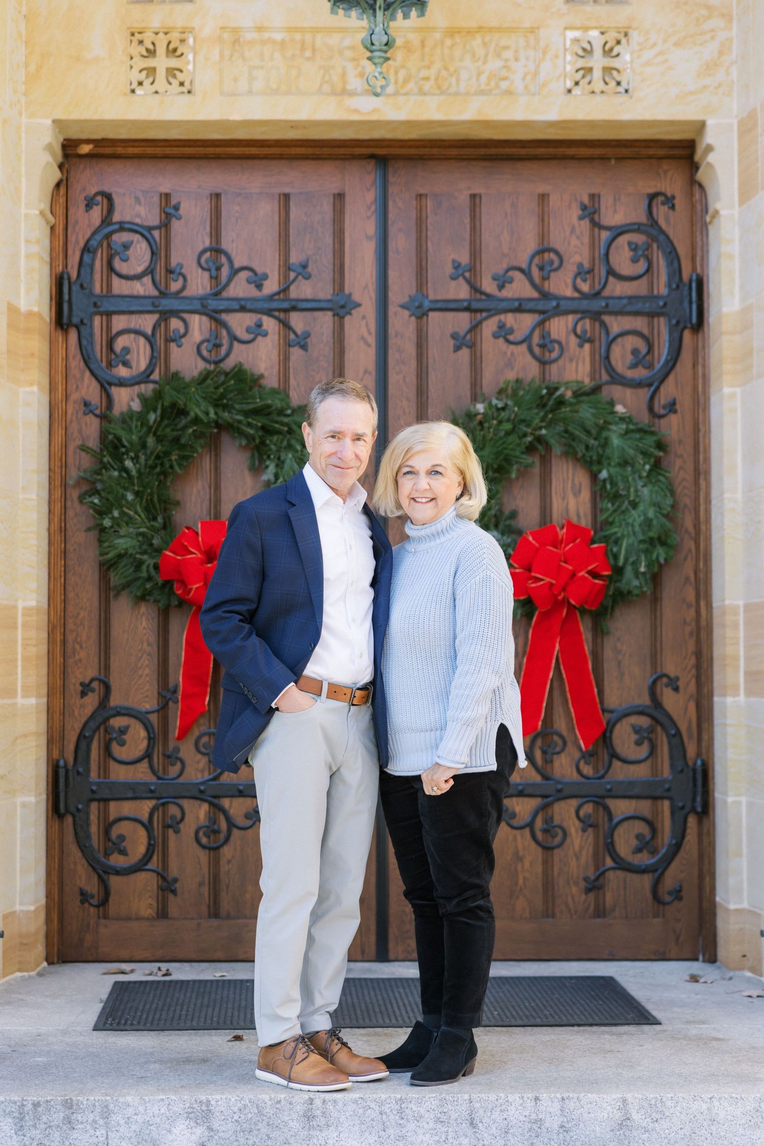Decorative wooden double doors with ornate iron hardware and two Christmas wreaths adorned with red bows.