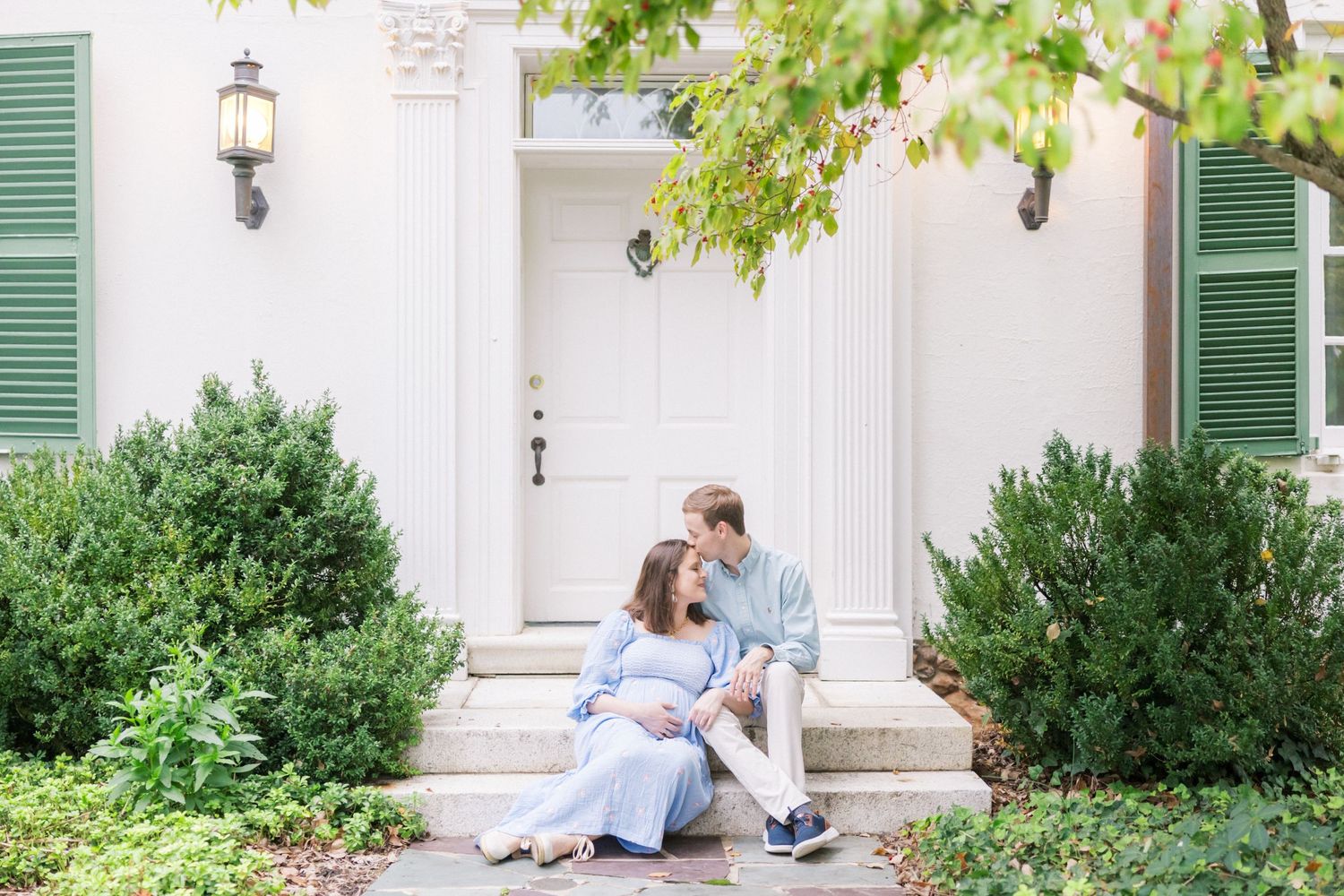 A couple sits on white steps outside a traditional home with green shutters and shrubs framing the entrance.