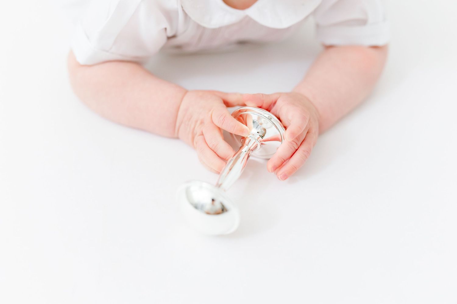 Baby in white outfit holding a small silver and white rattle toy with both hands on a white background.