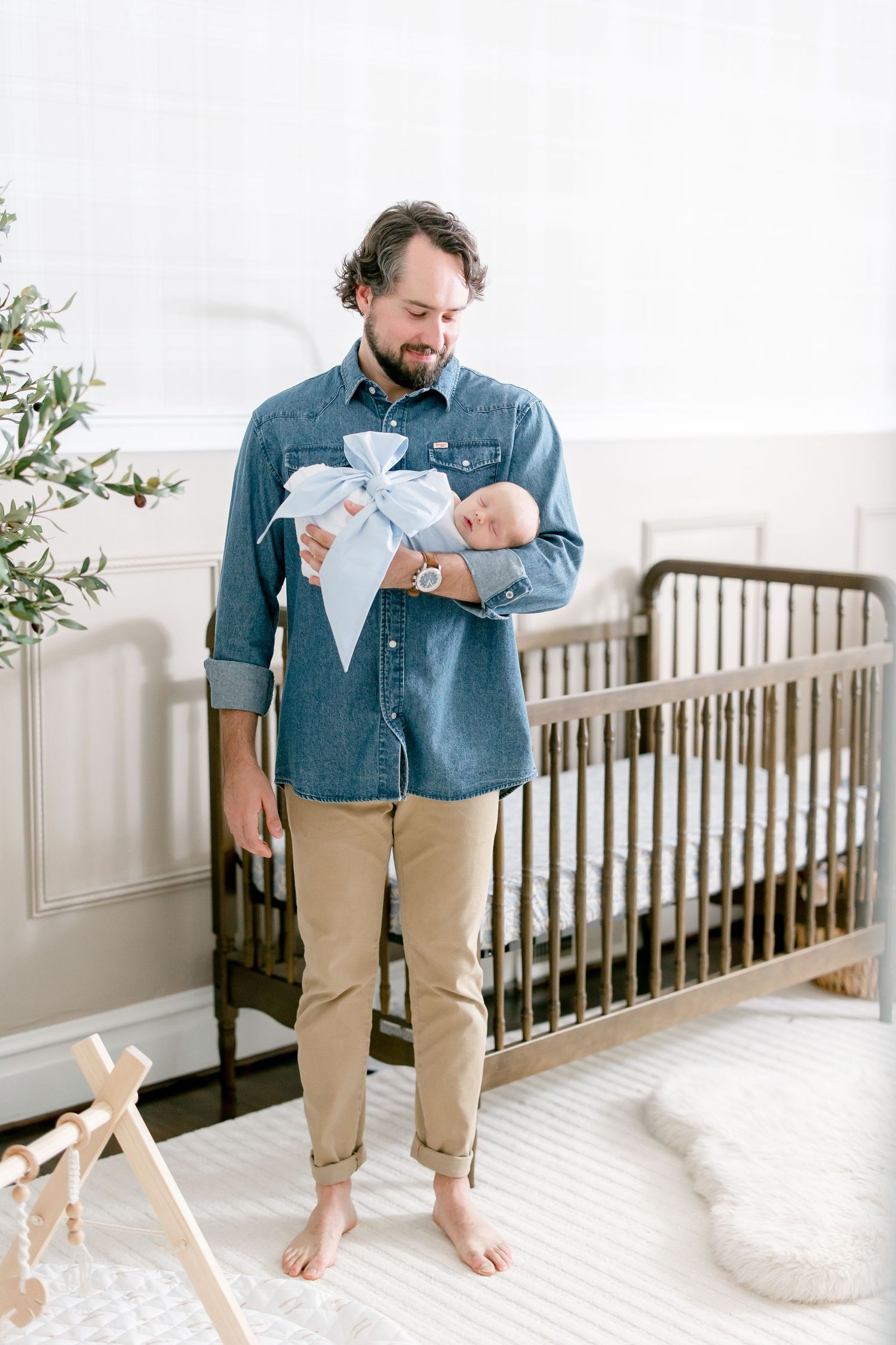 Parent in denim shirt and khakis holds and comforts a small baby in a bright white nursery.