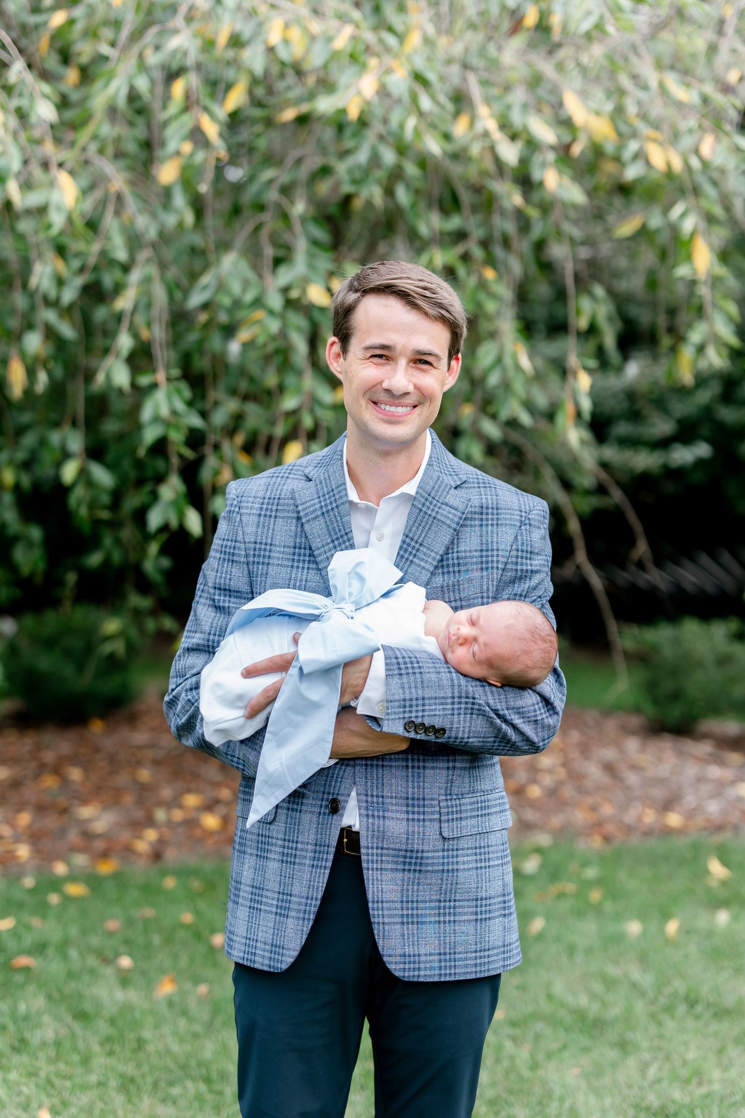 Man in plaid jacket holding baby against lush garden backdrop.