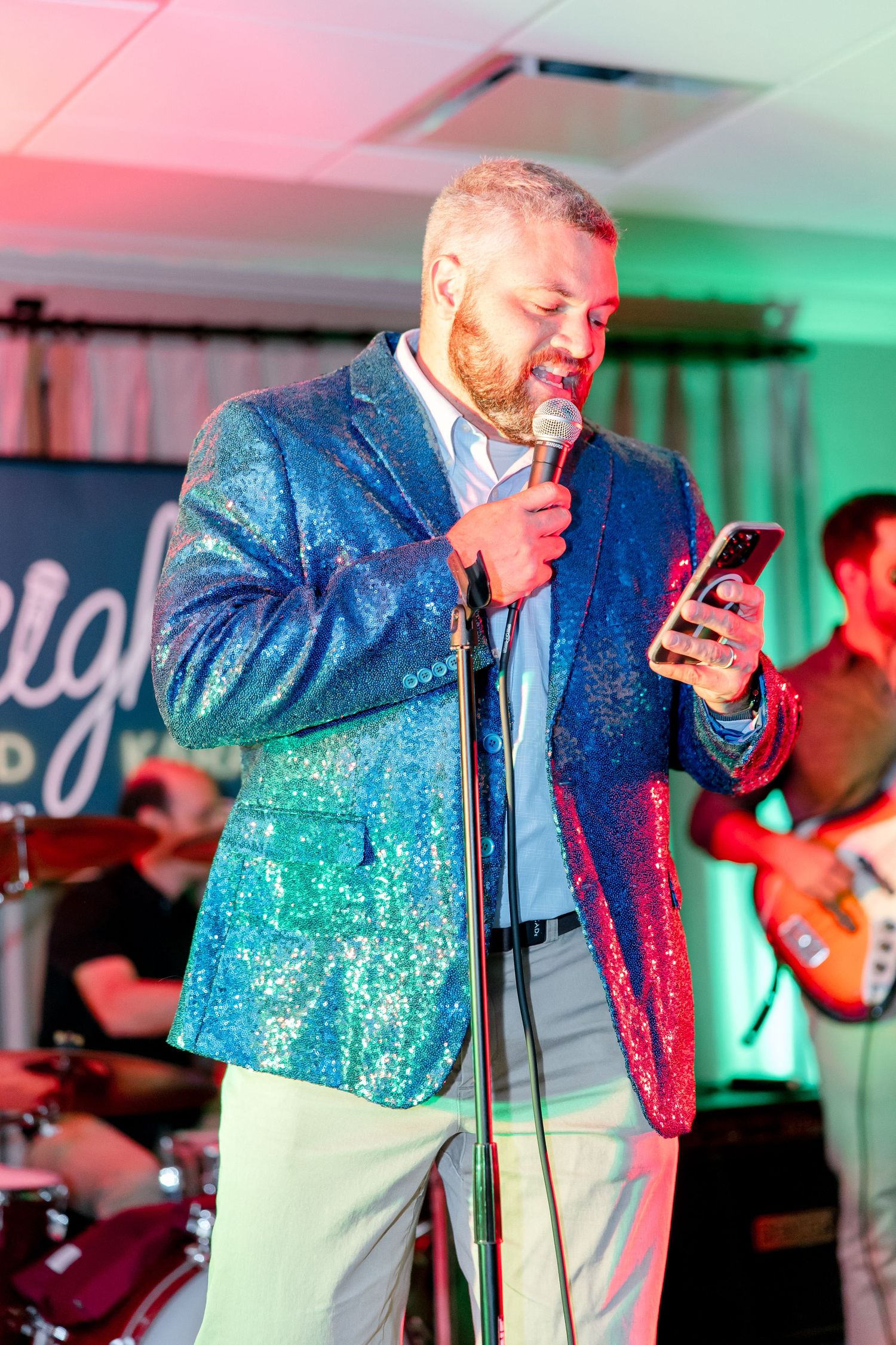 Performer in sparkly blue jacket sings into microphone while checking phone on colorfully lit stage.