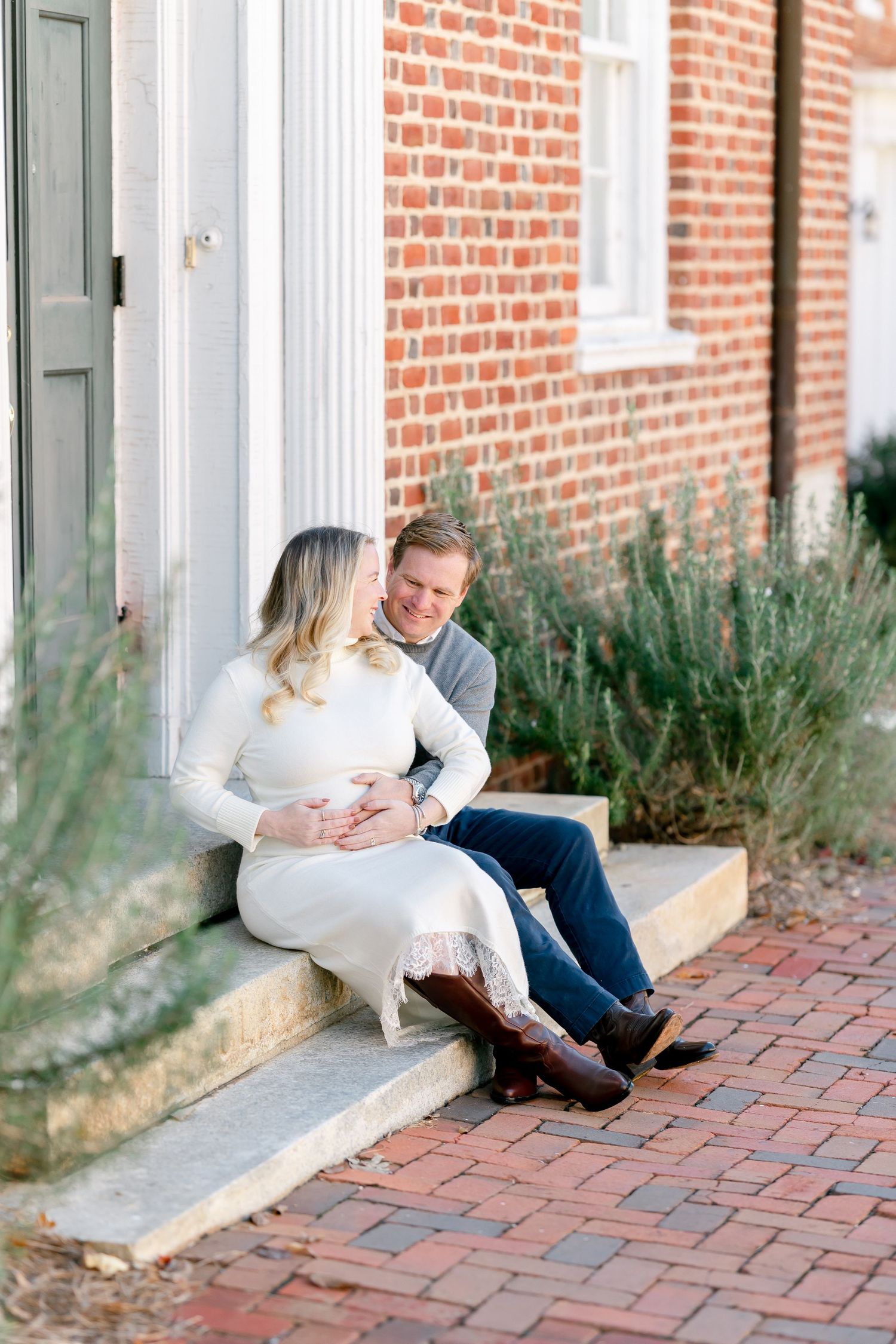 Smiling couple in casual attire sit together on stone steps beside a red brick building, the woman cradling her baby bump.