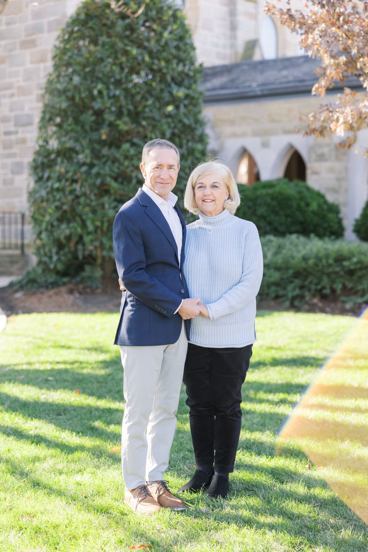 A couple stands together on a sunny lawn in front of a stone church with classic architecture.