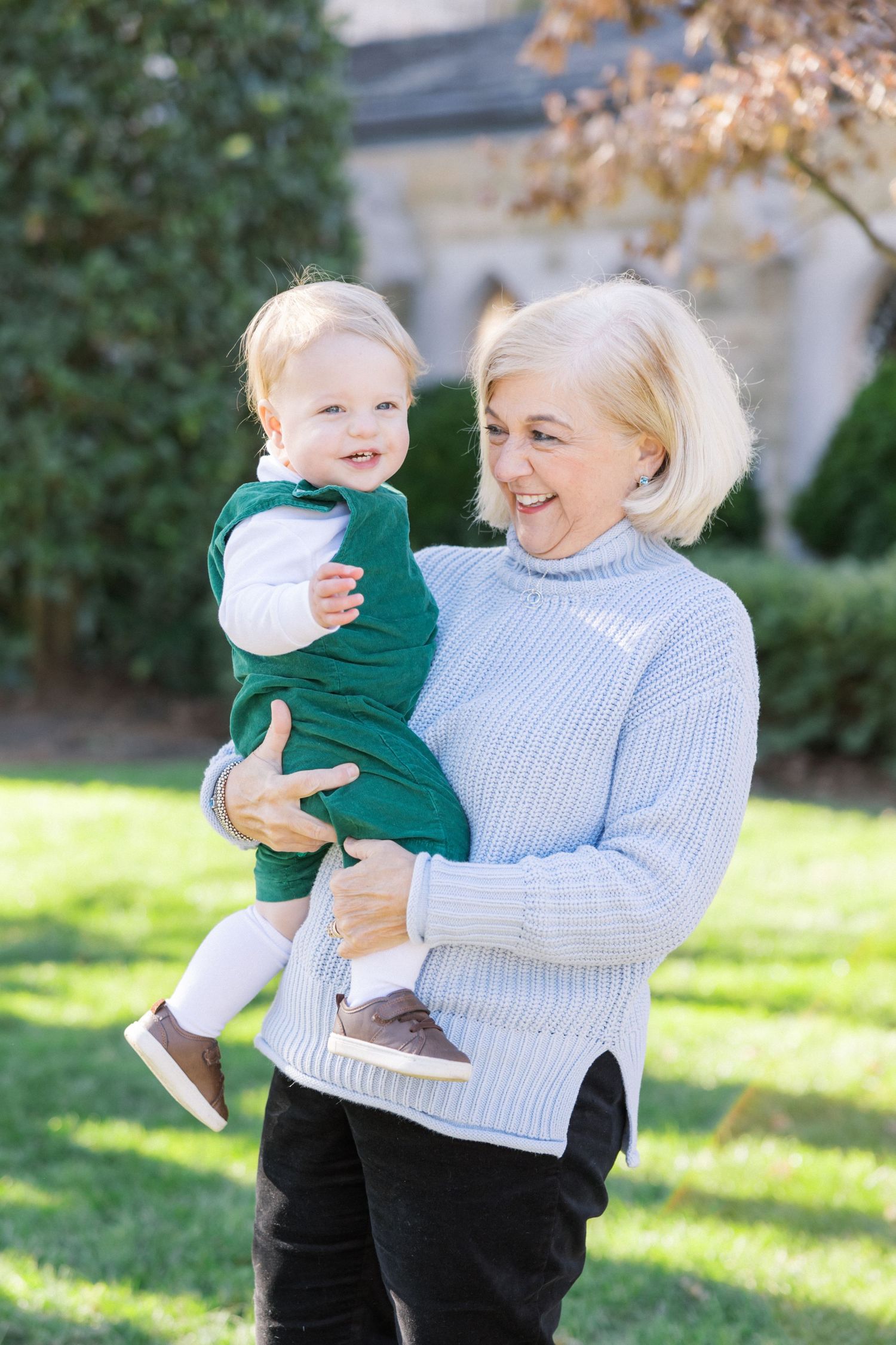 A person in a white sweater holds a small child wearing green outdoors on a sunny autumn day in front of a garden.