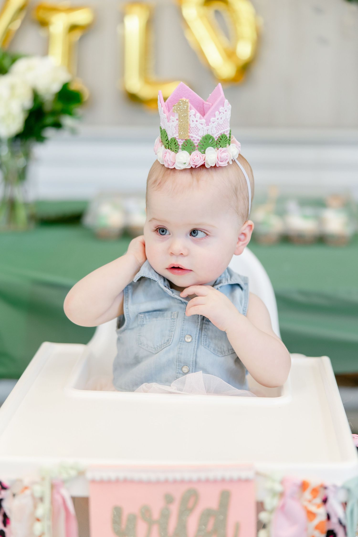 Baby in highchair wearing pink crown and grey outfit during first birthday celebration.