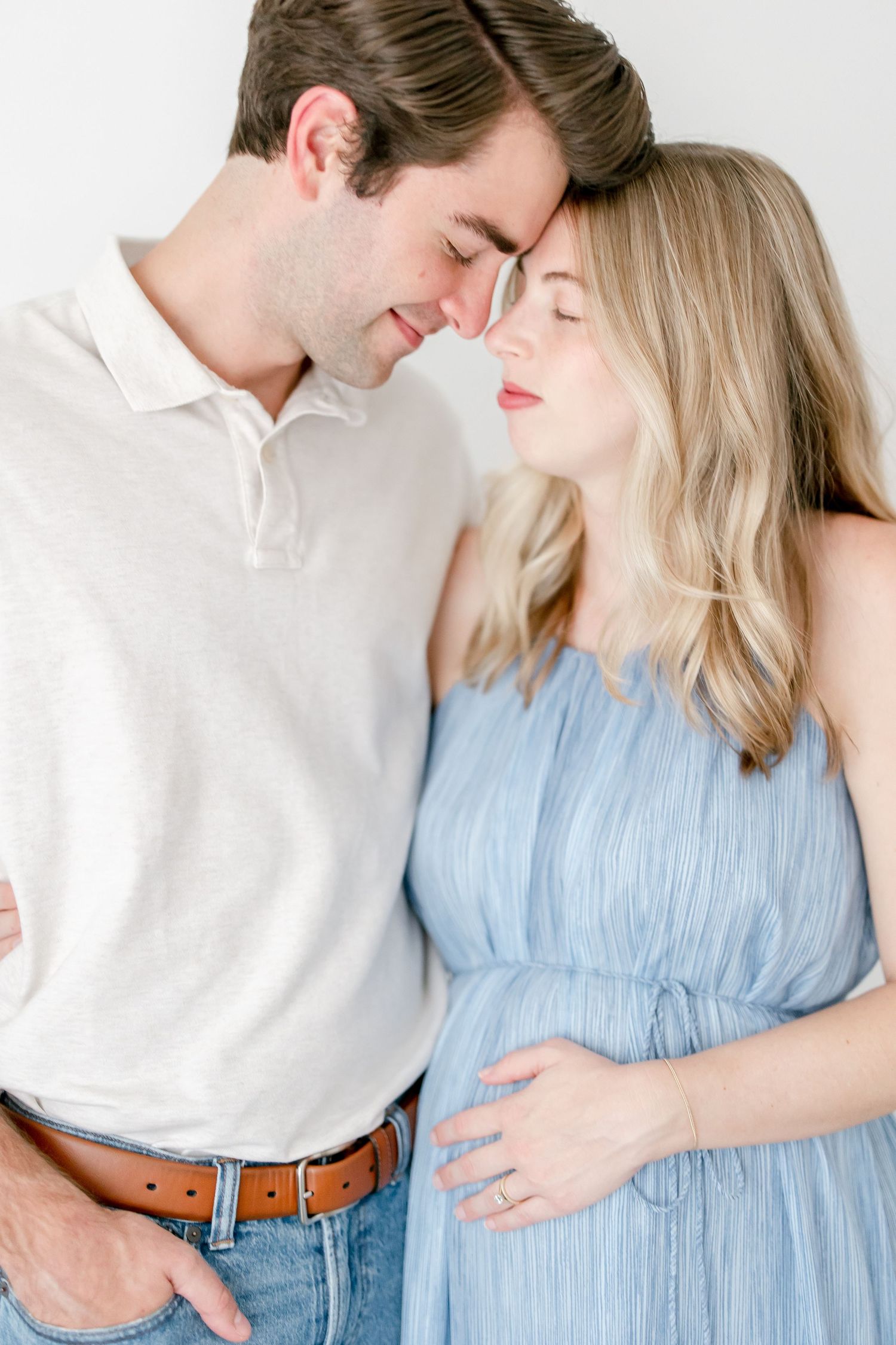 Couple in white shirt and blue dress pose romantically against white background in maternity photoshoot.