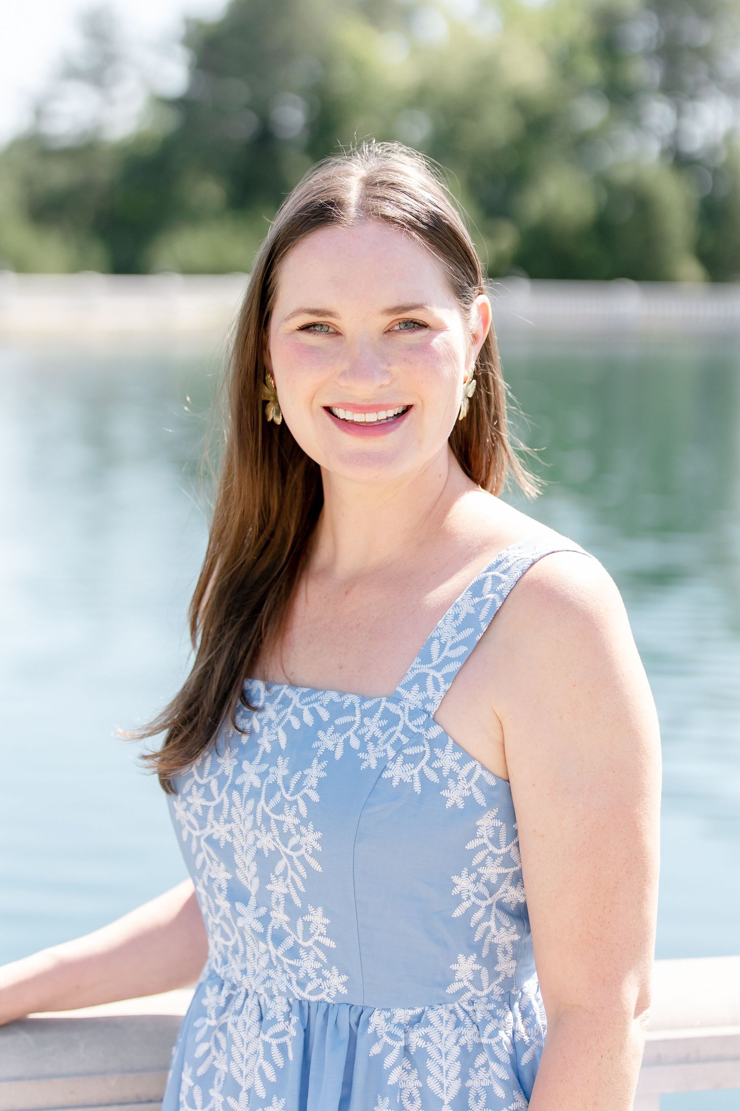 Woman in light blue floral sundress poses by lakeside on sunny summer day.