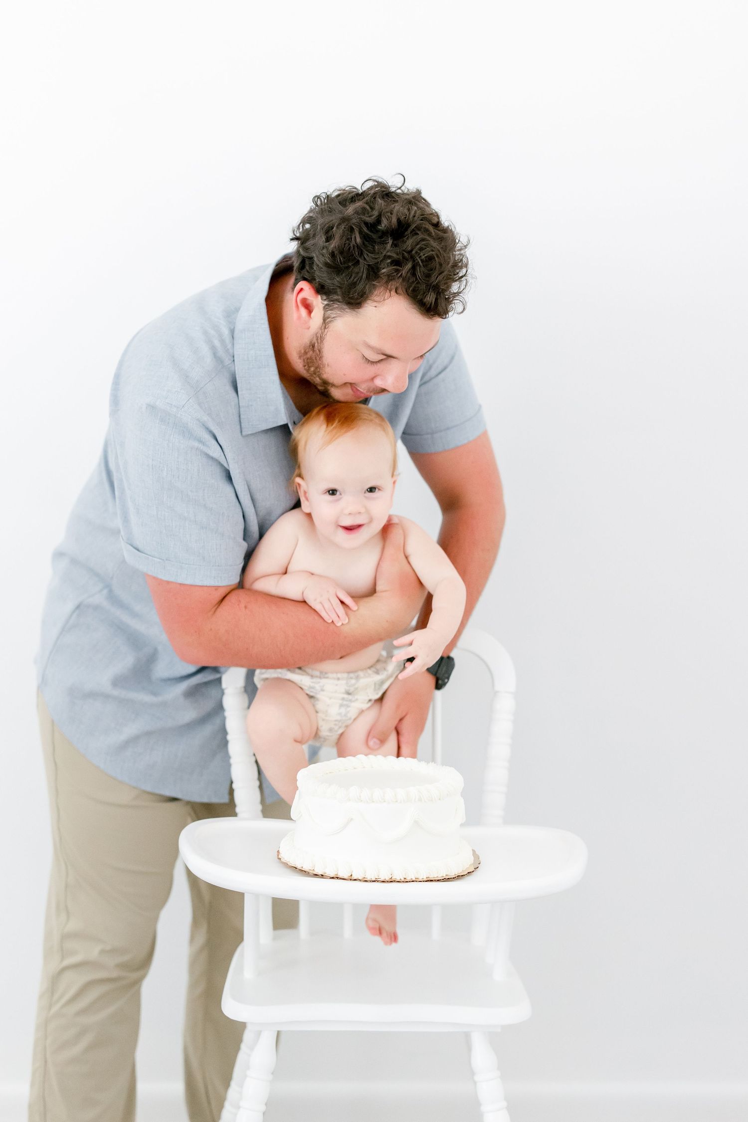 A parent in gray shirt and khaki pants helps their baby reach for a white birthday cake on a high chair.