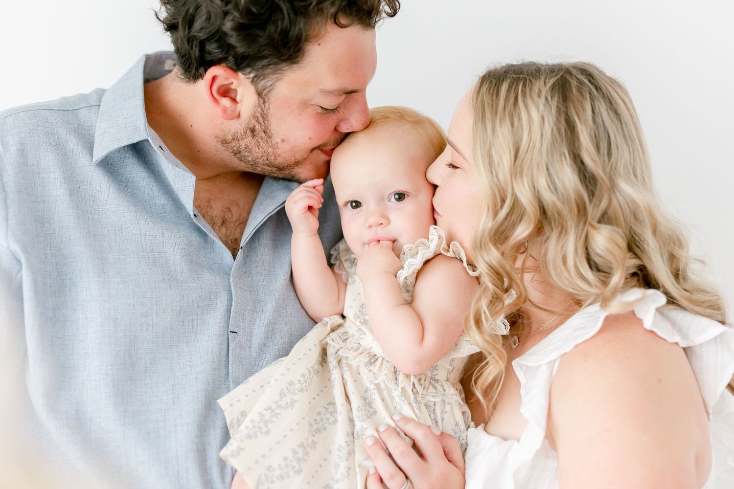 A couple lovingly embraces their baby wearing a cream-colored dress against a white background.