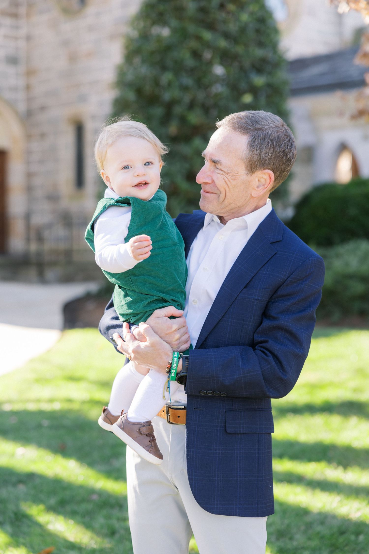 A well-dressed man in a navy blazer holds a small child wearing a green sweater outdoors near a stone building.